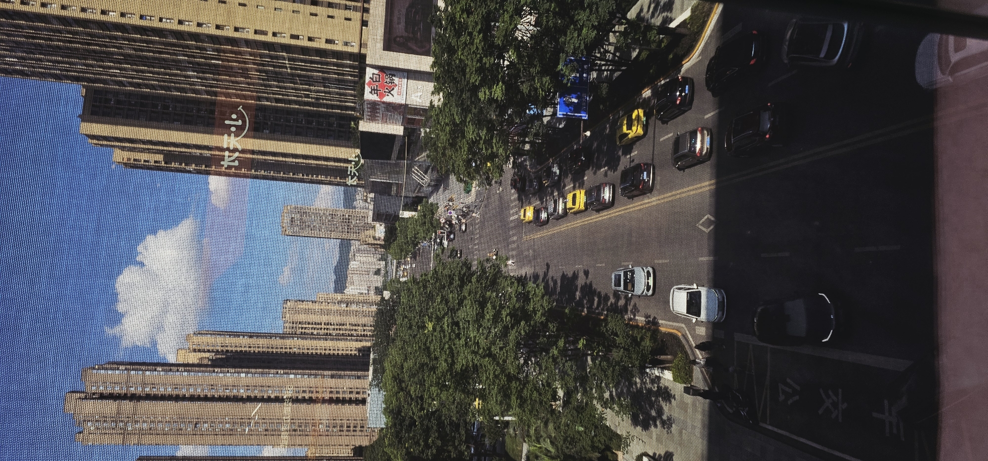 The image captures a view of a city street in Jiulongpo District, China, from an elevated perspective, likely from a window. Tall, modern apartment buildings dominate the skyline, characterized by their light-colored facades and numerous windows. The sky is blue with scattered white clouds, suggesting a clear day. A wide, paved road runs through the center of the frame, lined with lush green trees on either side, casting shadows on the asphalt. A steady flow of traffic is visible, with various cars, including sedans and what appear to be taxis, driving in both directions. Some cars are parked along the roadside. Pedestrians are also present, walking on sidewalks and crossing the street at designated areas. The scene appears to be a typical urban environment with commercial and residential structures. There are also billboards and signage on the buildings, some with Chinese characters. The overall impression is one of a bustling city scene under daylight.