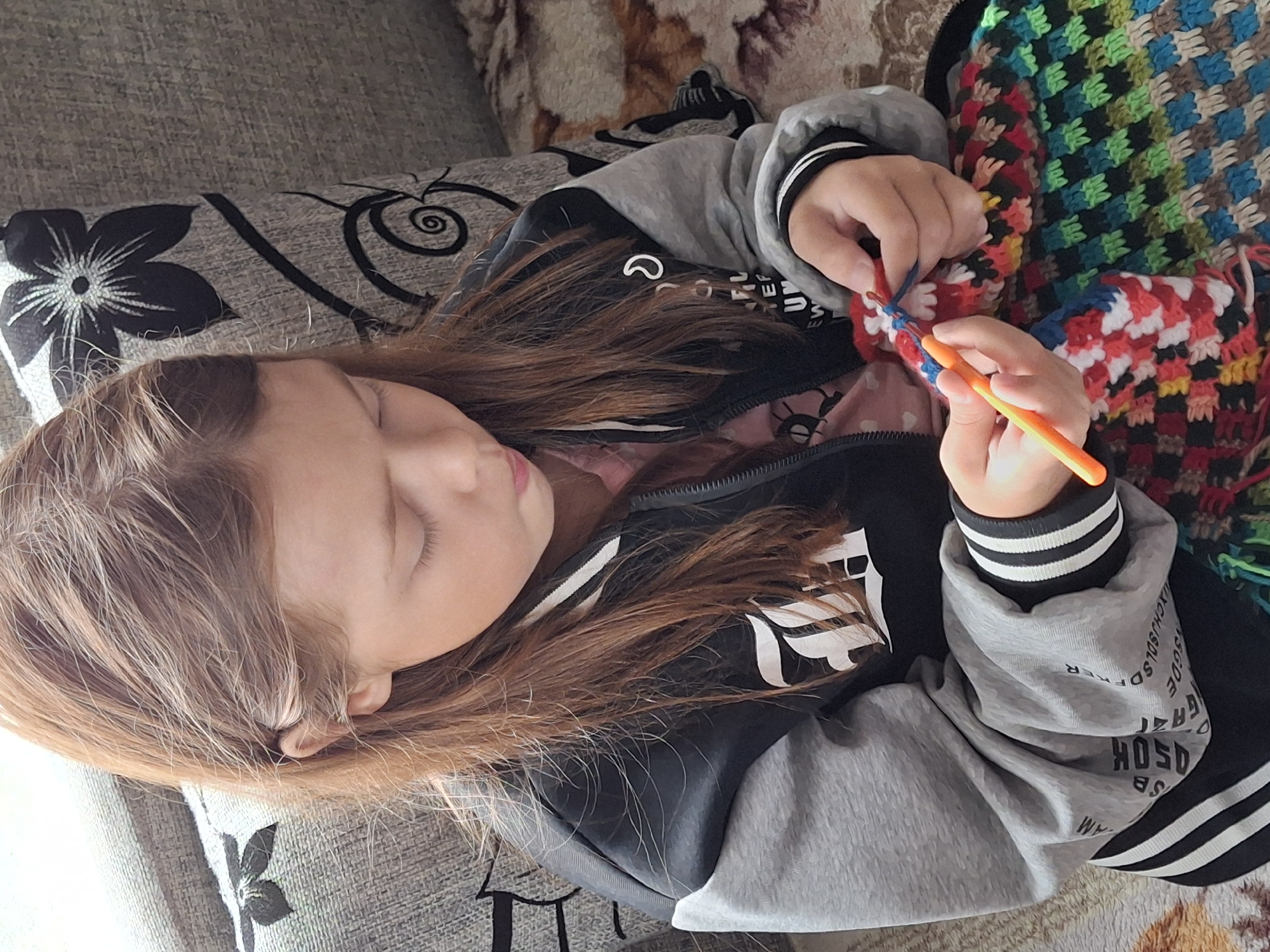 A young girl with long brown hair is sitting on a sofa and crocheting. She is wearing a black and gray jacket with white stripes on the cuffs and a pink shirt underneath. Her hands are focused on the colorful crocheted fabric, holding an orange crochet hook and blue yarn. The sofa is upholstered in a textured fabric with a floral pattern. The background is slightly blurred, showing more of the sofa and a patterned throw blanket. The scene suggests a moment of quiet activity, likely in a home environment in Plishkino, Russia.