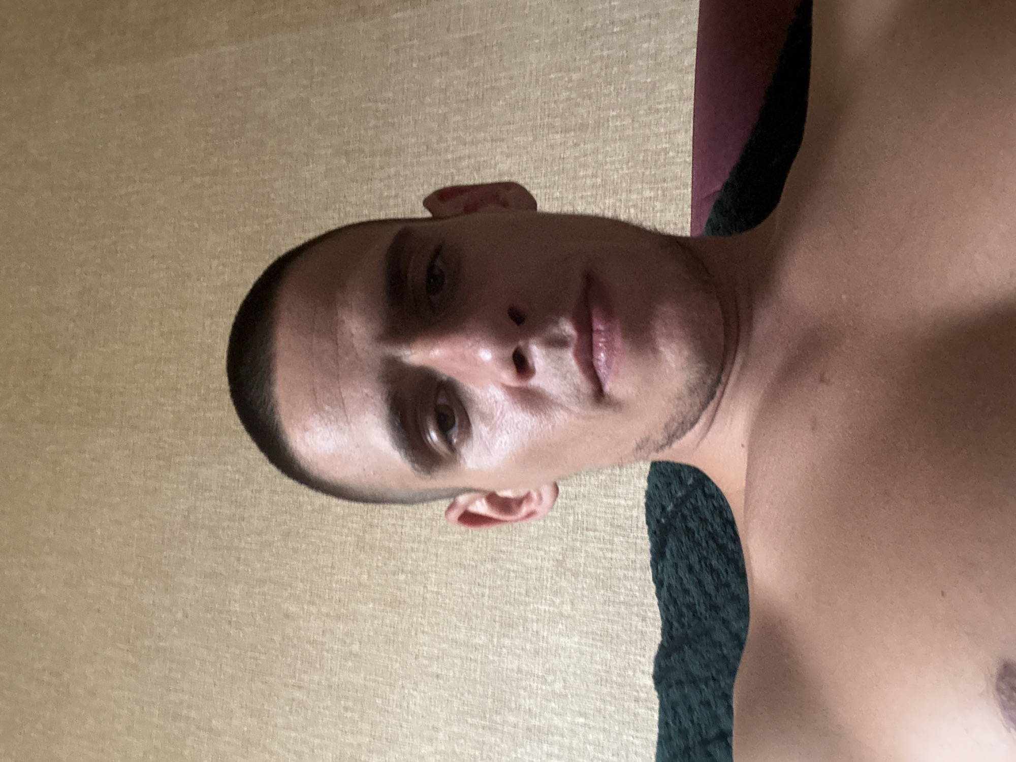 The image is a selfie of a man with a buzz cut. He is shirtless and looking directly at the camera. He is sitting on a piece of furniture, possibly a couch, covered with a dark green textured material. Behind him, the wall has a textured, light brown wallpaper. The lighting suggests an indoor setting, likely a residential space. The man's features are clearly visible, including his eyes, nose, and mouth. His shoulders and upper chest are also visible. There is no indication of the location being in Sofia, Bulgaria.