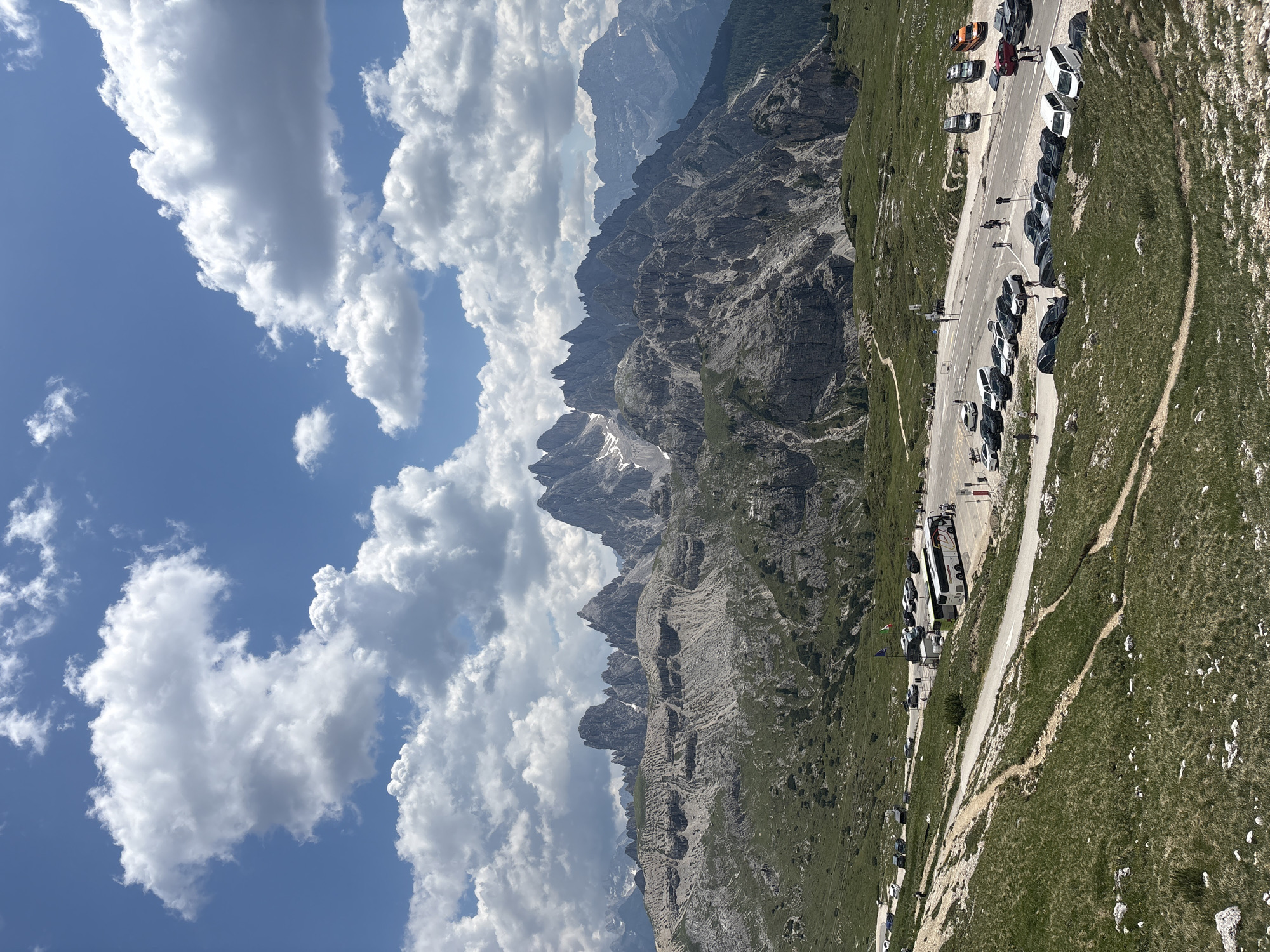 A scenic view of the Dolomites mountains in Italy. The foreground is a grassy slope with a dirt path winding through it. In the middle ground, there is a paved road with a parking lot filled with cars and a tour bus. People are seen walking around the parking lot. The background is dominated by the rugged, rocky peaks of the Dolomites, with a blue sky and fluffy white clouds overhead. The location is Tre Cime di Lavaredo, a national park in the Dolomites.