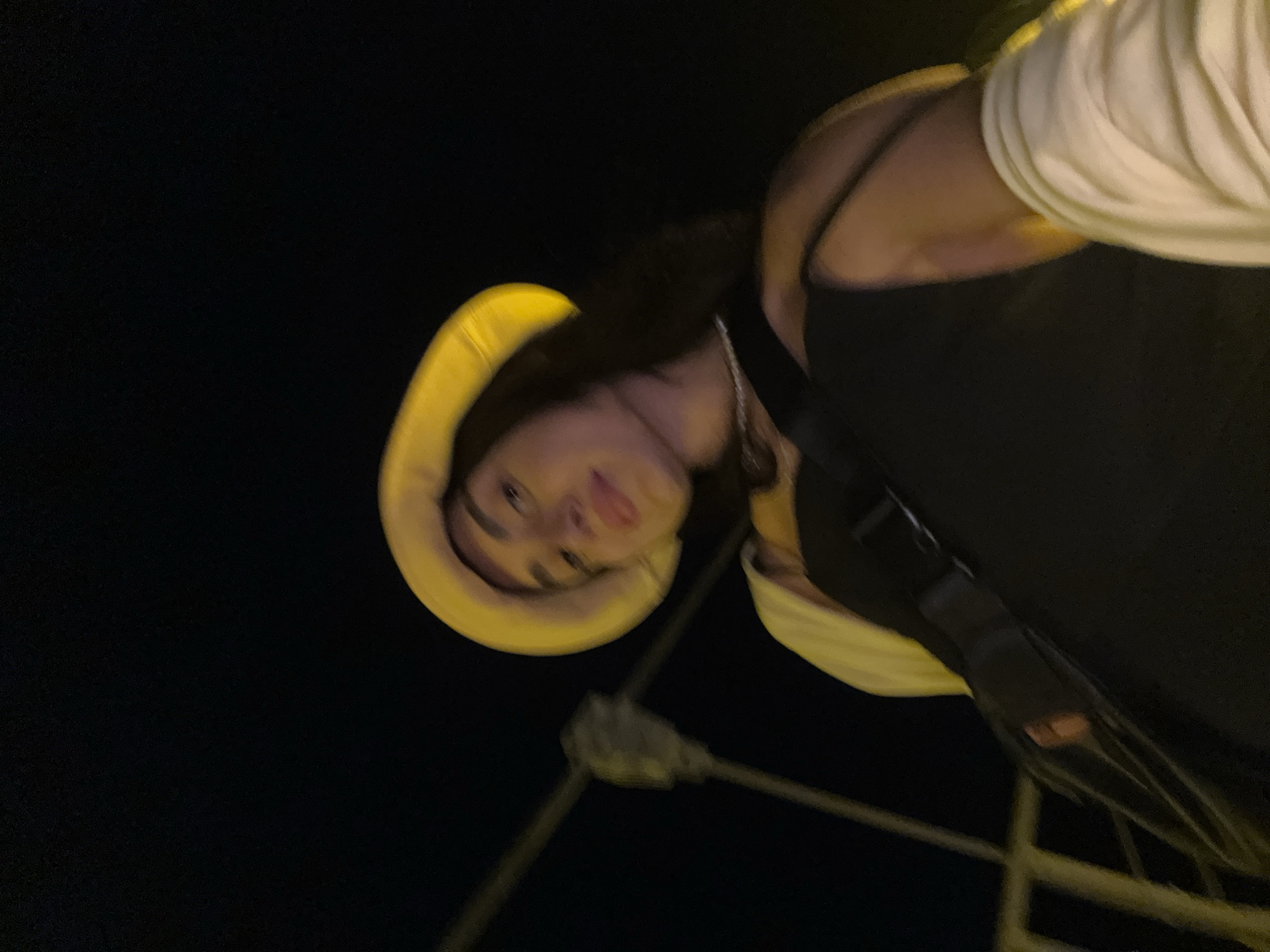 A young woman wearing a yellow bucket hat and a white cardigan over a black dress stands outdoors at night, appearing to be on a bridge in Poti, Georgia. The camera angle is from below, looking up at the woman as she looks down towards the camera. She has dark hair and is wearing a pearl necklace. A dark bag is slung across her chest. The background is dark, with some blurred streetlights and railings visible.