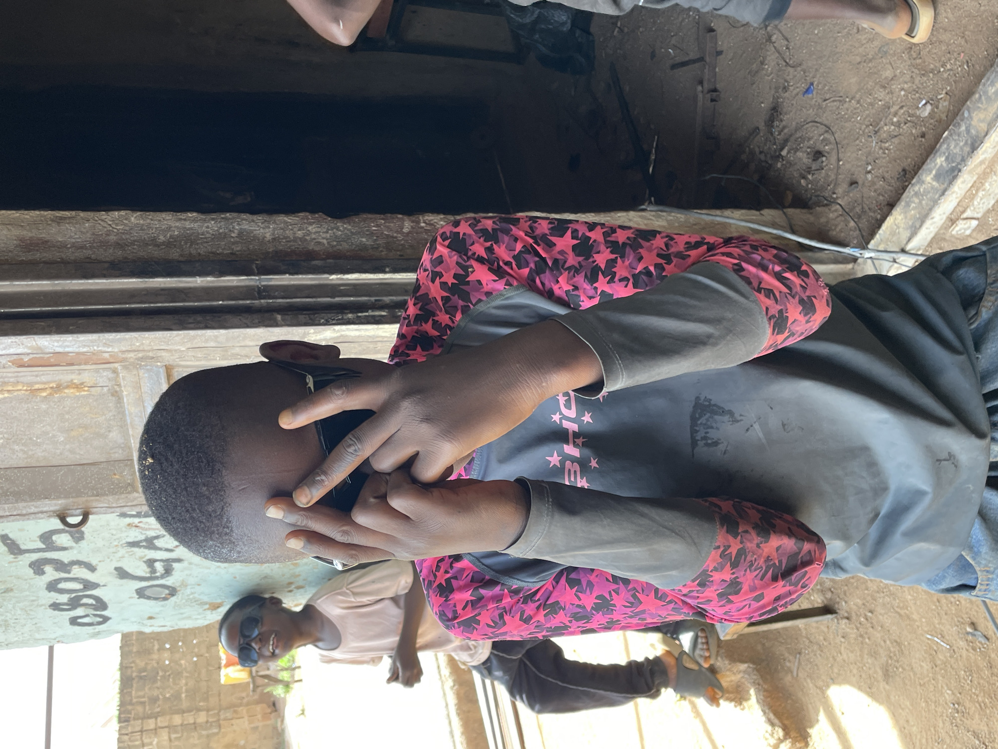 The image shows a young person indoors, possibly in a workshop in the city of Jos, Nigeria. The individual is wearing a long-sleeved shirt with a star pattern and is holding a phone to their ear. Another person wearing sunglasses is visible in the background. The floor appears to be concrete, with various tools and materials scattered around. A dark opening in the background leads to another space. Part of a painted sign or door is visible on the left.