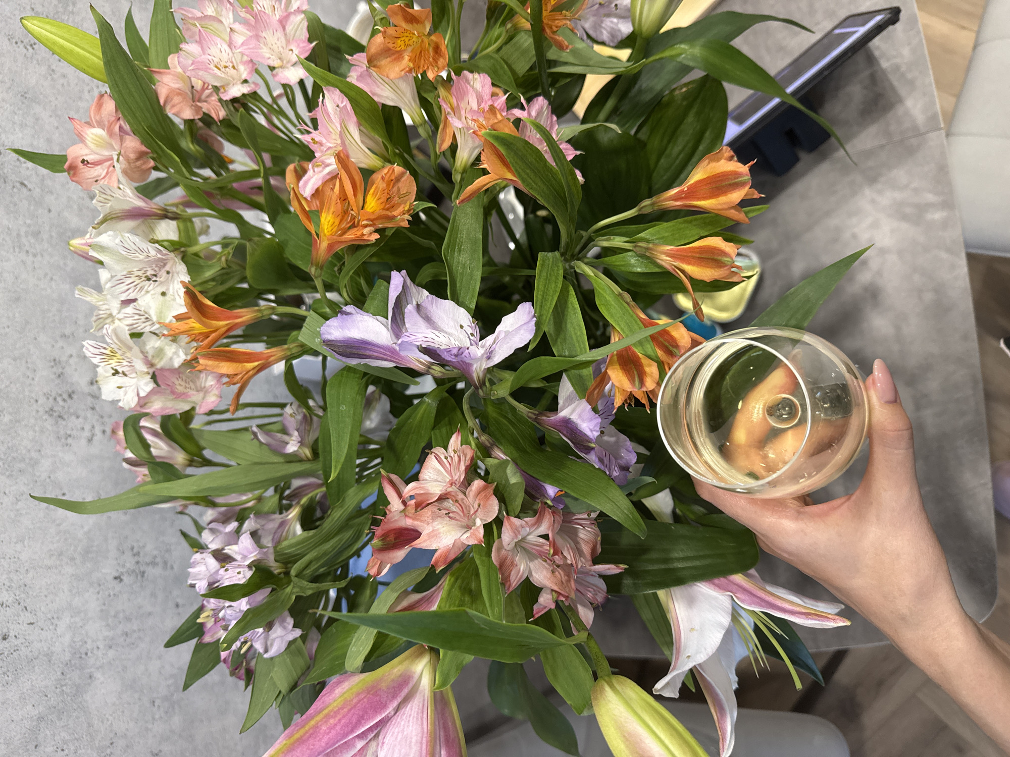 A high-angle shot shows a bouquet of mixed flowers, including orange, pink, white, and purple Alstroemeria and pink and white lilies, sitting on a gray surface. A hand holding a glass of clear liquid is partially visible on the right side of the frame. A smartphone in a dark case rests on the surface behind the flowers. A light-colored upholstered object is partially visible in the background. The location is Unknown, Unknown.