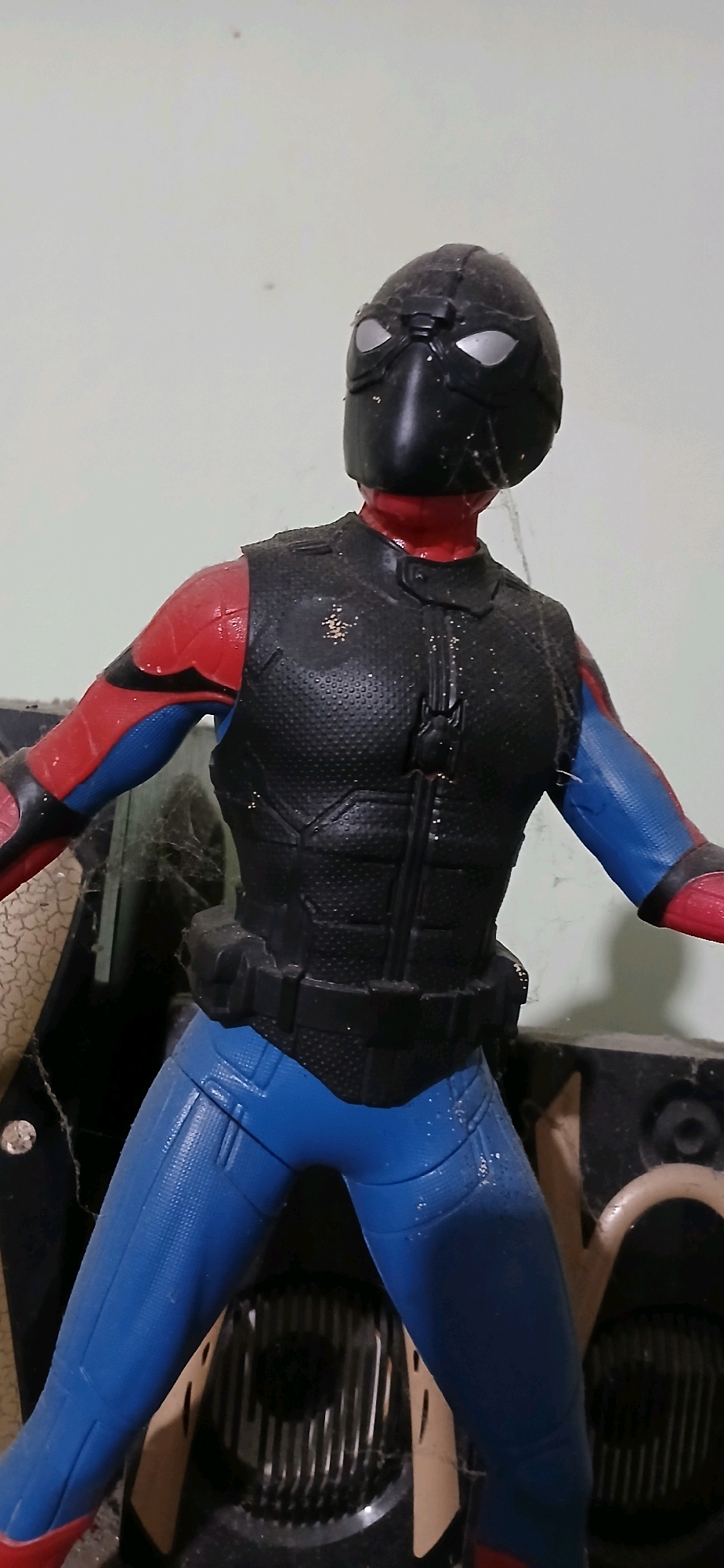 A toy figurine of Spider-Man is posed in a standing position against a plain, light-colored wall. The figurine is wearing a black mask with silver lenses, a red undersuit on its torso and arms, a black textured vest over the torso, and blue pants. The figurine appears dusty and has a few strands of cobweb on it, particularly around the vest and arms. In the background, partially obscured, are dark-colored objects that resemble electronic equipment or speakers, and a beige-colored surface with a textured pattern. There are some dust particles and potential cobwebs visible on the objects in the background as well. The lighting suggests an indoor setting. The location is specified as Sutawinangun, Indonesia.