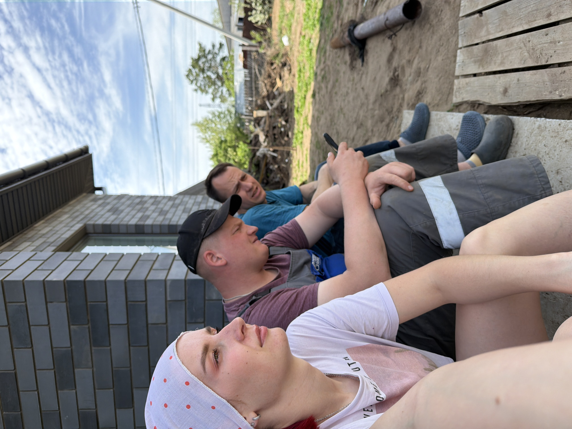 Three individuals are reclining on a concrete surface adjacent to a building with dark brickwork, likely located in Chaikovsky, Russia, based on contextual clues. The foreground features a person wearing a white t-shirt and a white headscarf with red polka dots. Slightly behind, a person in a burgundy t-shirt and dark overalls with a black baseball cap is also reclining. Further back, a third person wearing a blue t-shirt sits while holding a phone in their hand. All three appear relaxed, and their legs are stretched out.
The setting includes a building, a patch of sand or dirt, and what appears to be a wooden structure. A pipe lies on the sand. Green vegetation and a partly cloudy sky are visible in the background. The image is taken from a low angle, possibly from the ground.