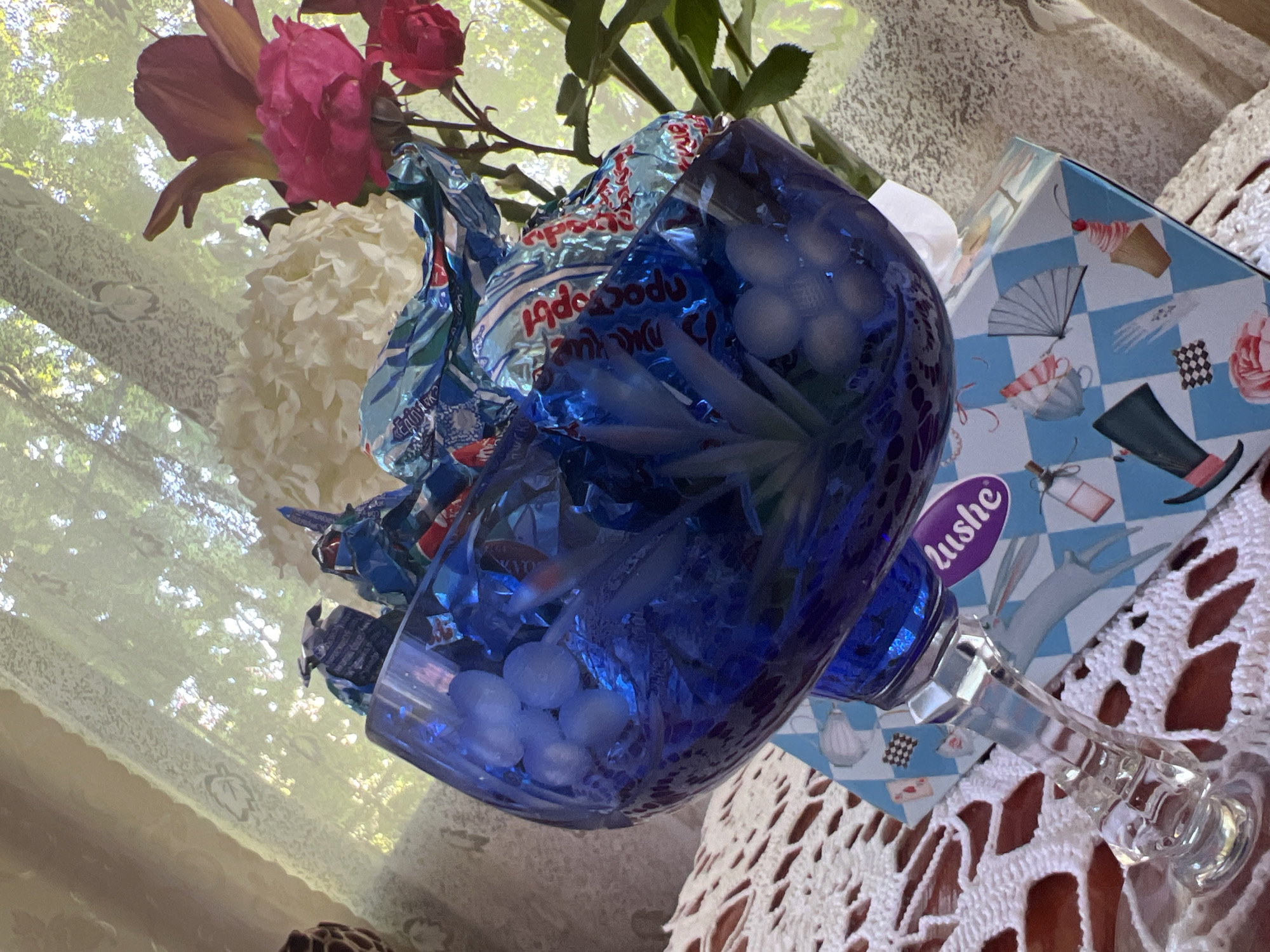 The image shows a blue glass vase filled with chocolates wrapped in blue foil. The vase is placed on top of a blue and white checkered box that has Alice in Wonderland-themed illustrations. Behind the vase and flowers, a sheer curtain with a tree pattern is visible, and through the curtain, greenery and sunlight can be seen, suggesting an outdoor setting visible from an indoor window. The image appears to be taken in Lopatino, Russia, as indicated by the "Lushe" branding on the box, which is a Russian chocolate brand. The scene is a still life arrangement with flowers and candy.
