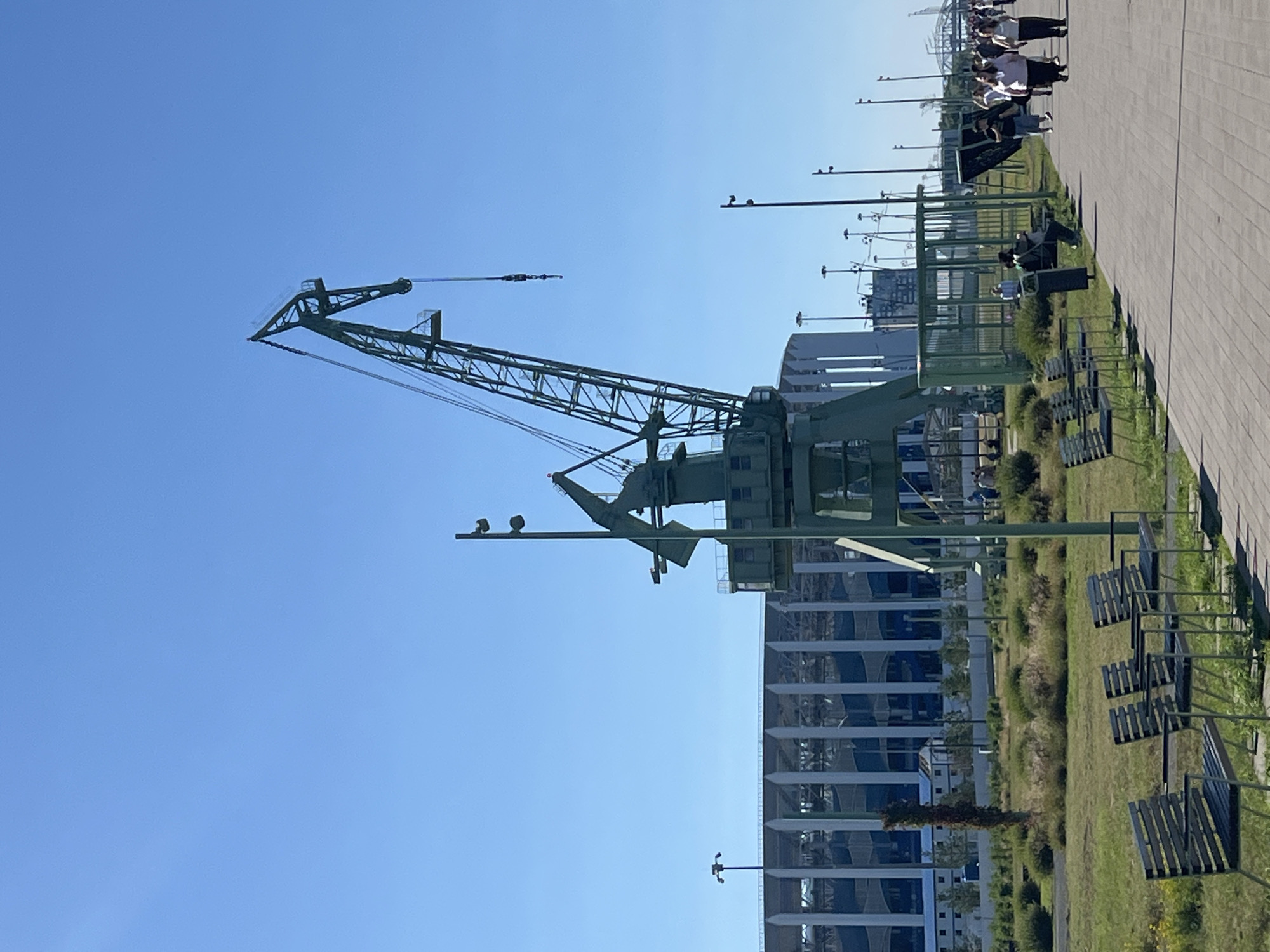 A large, green, industrial crane stands prominently in the foreground against a clear blue sky. The crane is a fixed gantry crane, with a tall, lattice-work boom extending upwards and to the right. The base of the crane appears to be a heavy, boxy structure. In the background, to the left of the crane, is a large, modern stadium with a white, slatted facade and a blue roof structure. The stadium is partially obscured by the crane and some vegetation.
In front of the stadium and to the right of the crane, a paved walkway runs horizontally across the image. People are walking along the walkway, mostly in groups. A row of dark-colored benches with slatted seats and backs are positioned along the edge of the walkway, facing a grassy area with scattered bushes. There are also several tall, slender lampposts visible throughout the scene. A green, caged structure, possibly a shelter or display area, is situated near the walkway.
The overall environment appears to be a public space or park adjacent to a waterfront or industrial area, given the presence of the crane and stadium. The image seems to have been taken on a sunny day. The location is identified as Nizhny Novgorod, Russia, likely near the Nizhny Novgorod Stadium, which is visible in the background.