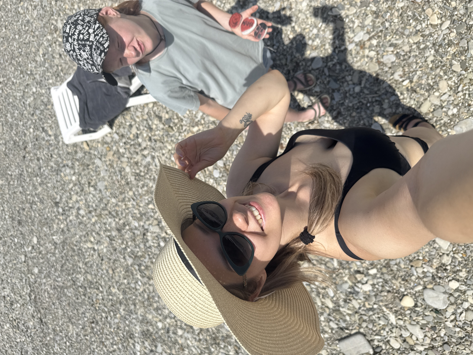 An overhead selfie captures two individuals on a pebble beach in Nebug, Russia, under bright daylight conditions.
In the immediate foreground, an adult female is positioned, her head and upper torso angled upwards towards the camera, which she is holding. She possesses blonde hair, styled with a small ponytail secured by a black elastic band visible on her left side. She wears a wide-brimmed, light-colored straw hat with a dark band, and dark, cat-eye style sunglasses with a small metallic emblem on the temple. Her lips are parted in a smile, revealing teeth. Her left forearm, partially visible, displays a small black, stylized tattoo. She is wearing a black bikini top and dark sandals. Her right hand is lightly touching the brim of her hat.
Behind and to the right of the foreground individual, a younger person, appearing to be a male child or teenager, stands on the pebbled ground. He has reddish-brown hair and wears a black and white patterned baseball cap with visible text including "SPIDER-MAN." He is wearing a light greenish-grey short-sleeved t-shirt, dark shorts, and dark sandals. A dark, thin choker-style necklace is visible around his neck. His facial expression is somewhat contorted, with his eyes appearing partially closed or squinting. His left palm is open, displaying five small, heart-shaped objects: three red, one white, and one black, each with a distinct design.
The ground is composed entirely of small, varied grey and white pebbles and stones. In the mid-background, behind the younger person, a white plastic reclining beach chair is visible, draped with a dark fabric item. Strong, distinct shadows are cast on the pebbled surface, indicating direct sunlight from a high angle.