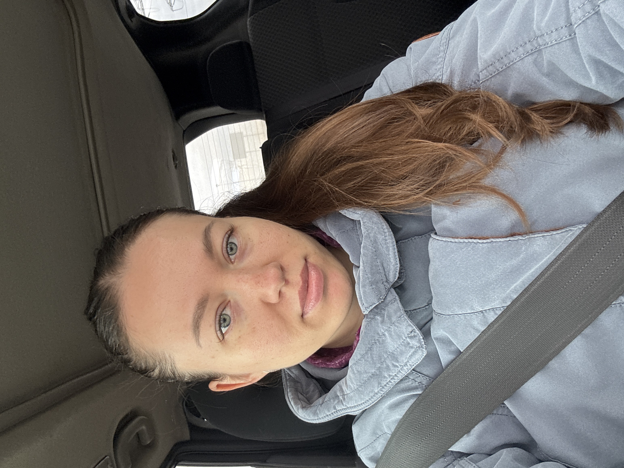 A woman with light skin and brown hair is sitting in a car, wearing a grey jacket and a seatbelt. The interior of the car is visible, including the seats and dashboard. Through the car window, a building is visible. There is no evidence to indicate the location is Lipaki, Russia.