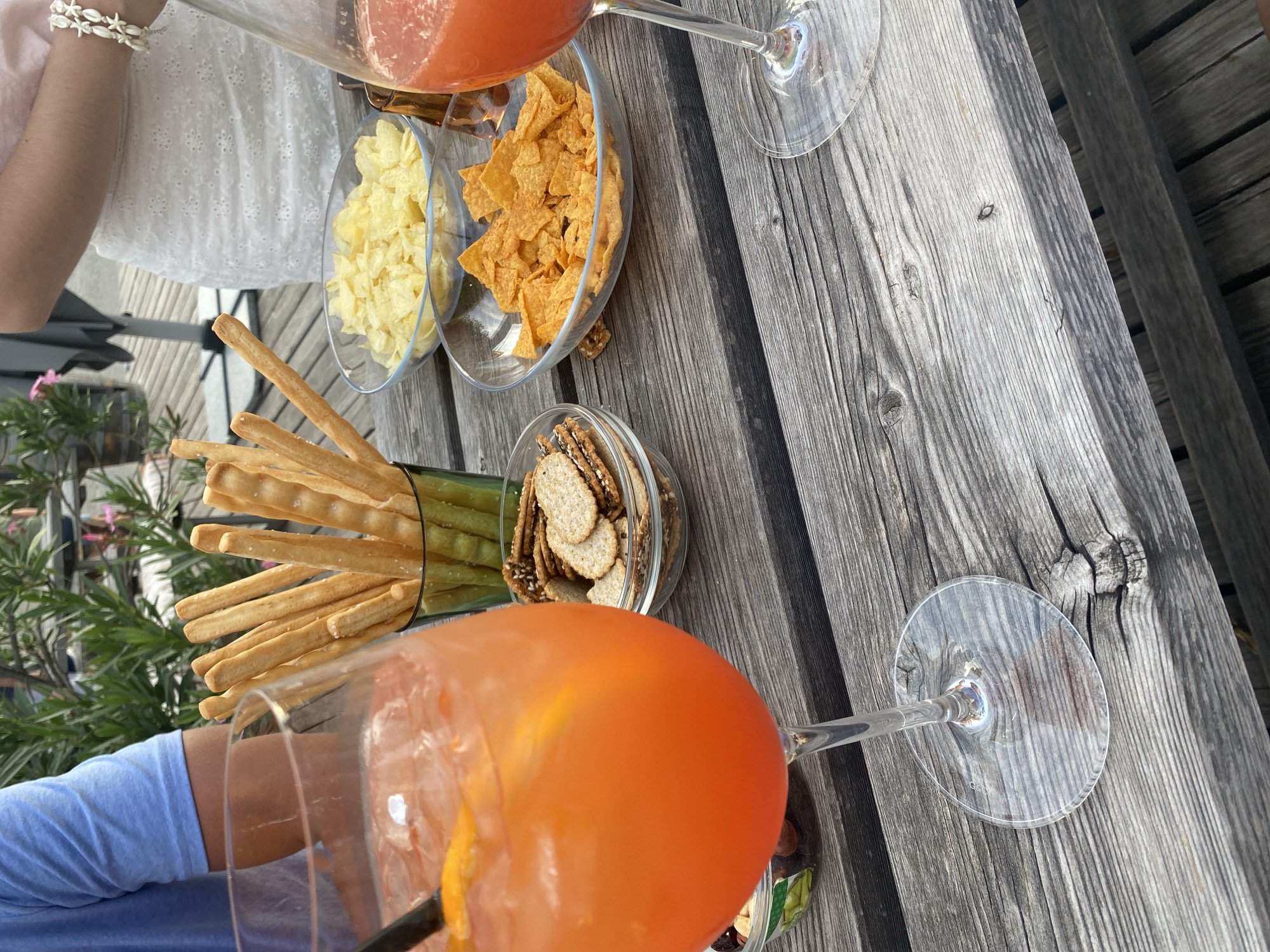 In Purbach, Austria, an outdoor table setting is captured on a weathered wooden surface. The composition features two stemmed glasses, one containing an orange beverage with ice and a garnish of orange peel, and the other partially visible with a similar red-hued drink. Several bowls are arranged on the table: one holds an assortment of crackers, another contains potato chips, and a third is filled with orange tortilla chips. A green glass container holds several breadsticks. In the background, two individuals are partially visible; one on the left wears a blue t-shirt, and another on the right wears a white lace skirt and a pink top, adorned with a white shell bracelet. Lush green foliage forms a backdrop, suggesting an outdoor seating area, possibly a terrace or garden.