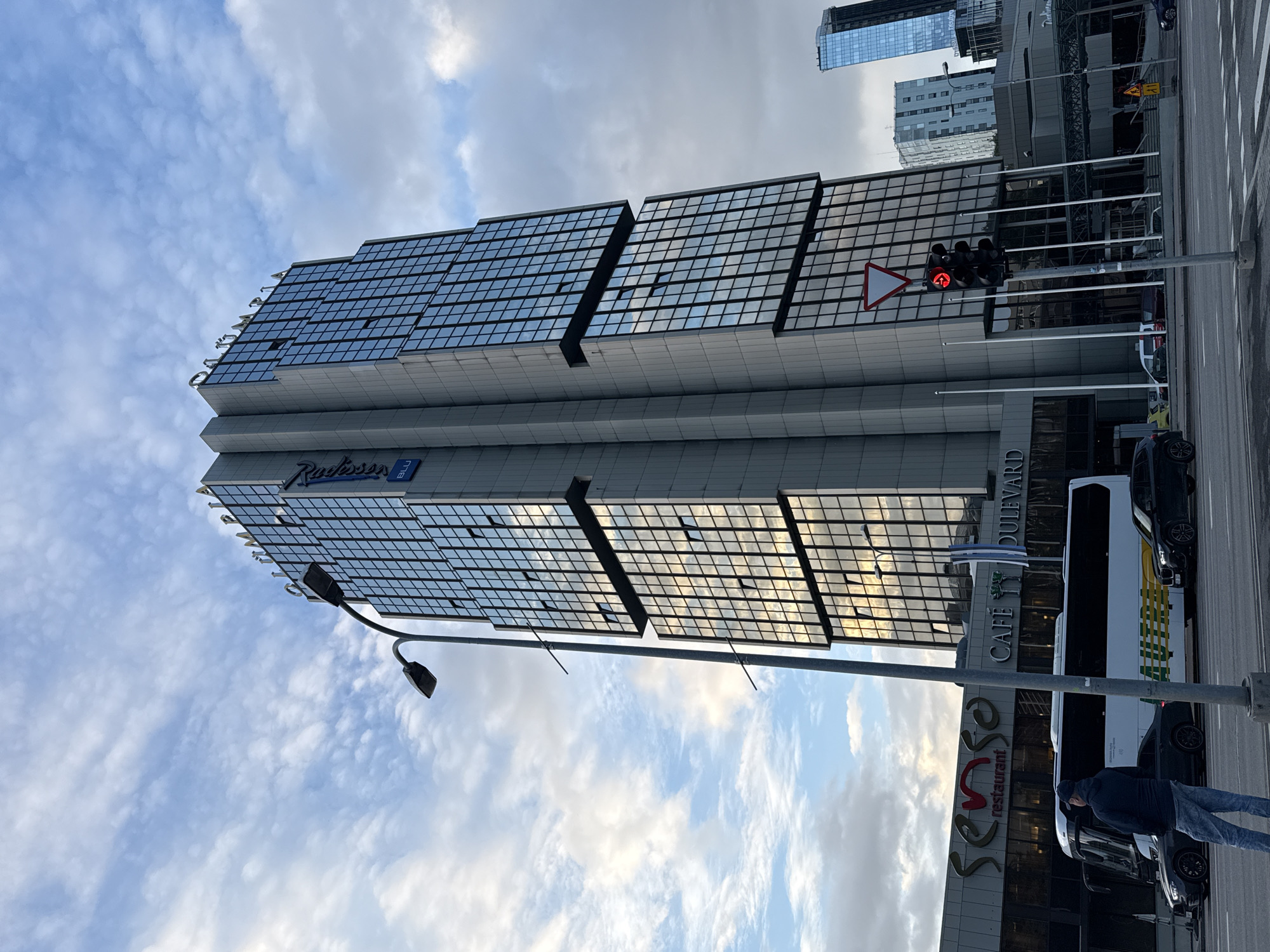 The image depicts a wide-angle view of the Radisson Blu Sky Hotel in Tallinn, Estonia, situated in the city center. The imposing glass-and-concrete skyscraper is the dominant feature, reflecting the cloudy sky and surrounding urban landscape. In the foreground, a street with a traffic light displaying a red arrow is visible, indicating traffic control. Several vehicles, including a bus, are parked or moving on the street. To the left of the hotel, a lower building houses a restaurant called "Senso" and a cafe named "Cafe Boulevard." The overall atmosphere is that of a metropolitan city during daylight hours, with a mix of modern architecture and typical urban elements.