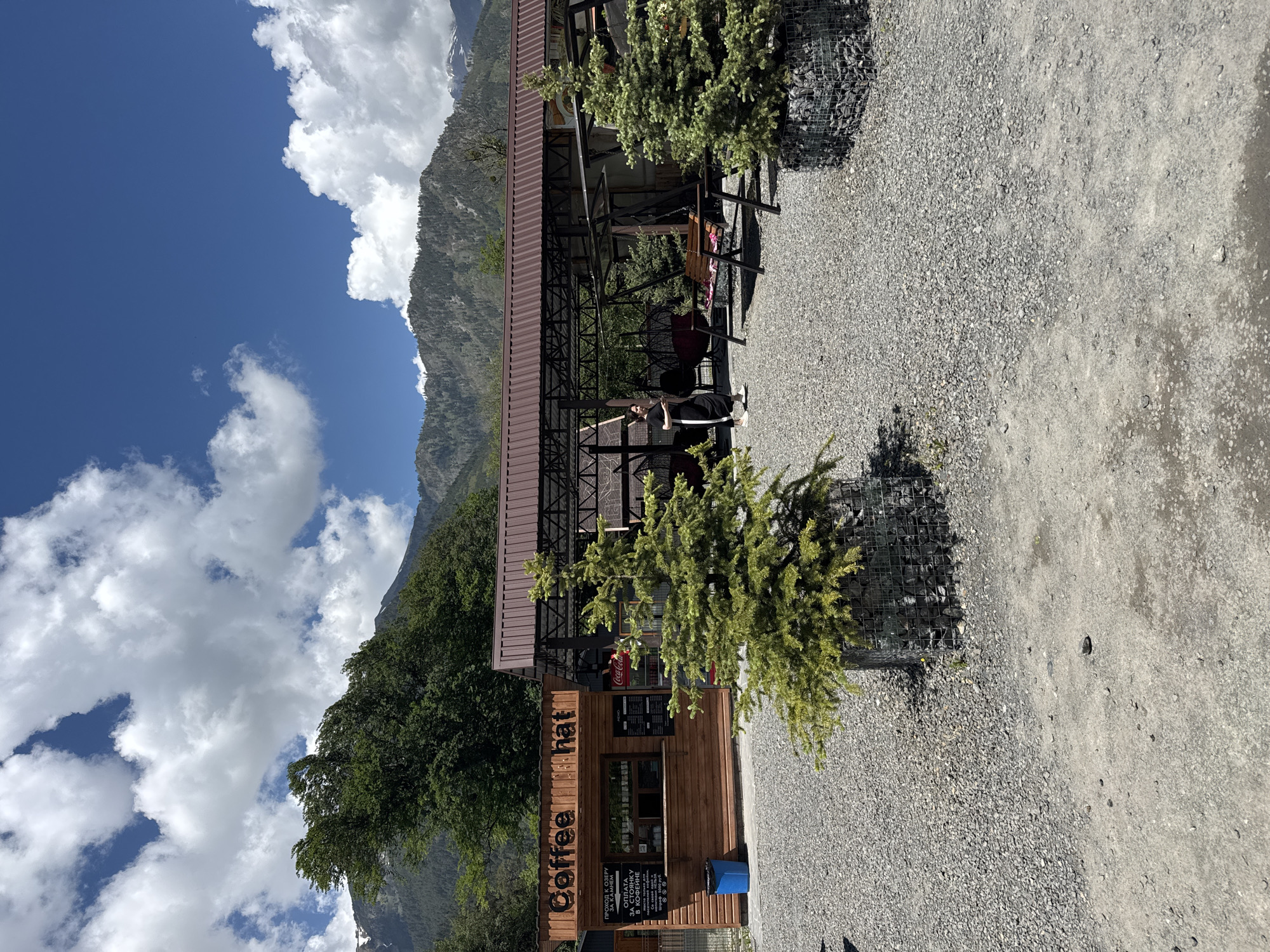 In the city of Teberda, Russia, a wooden coffee stand named "Coffee hat" is situated in an outdoor area. The stand has a counter and a menu board. To the right of the coffee stand is a covered seating area with tables and swings. A woman in athletic wear is standing near the seating area. The ground is covered with gravel, and there are several small evergreen trees in gabion planters. In the background, mountains with scattered trees and patches of snow are visible under a blue sky with white clouds.