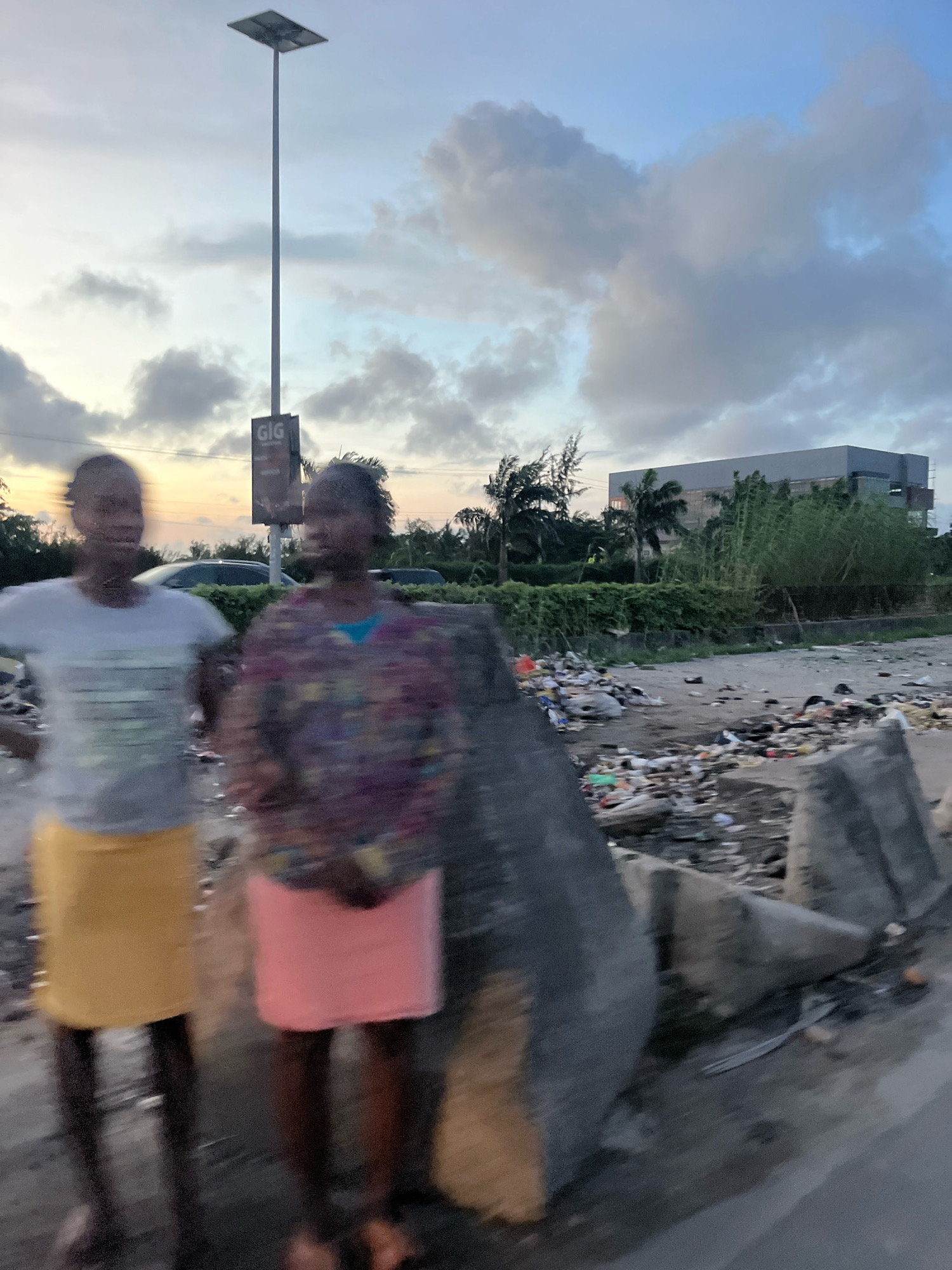 Two young women stand on a street in Ajah, Nigeria, amidst a scene of urban decay. The woman on the left wears a light-colored t-shirt and a yellow skirt, while the woman on the right is dressed in a patterned top and a pink skirt. Both are somewhat blurred due to motion. In the background, a tall lamppost with a solar panel is visible, along with a sign for "GIG" and a multi-story building. The ground is littered with significant amounts of garbage, including plastic bags and other debris. Palm trees and dense green foliage line the area behind the garbage, and the sky above is partly cloudy with the warm hues of twilight. The overall environment suggests a neglected roadside or public space.