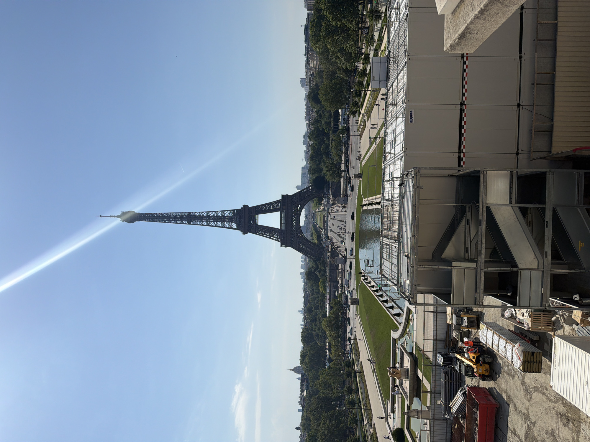 The image captures a view of the Eiffel Tower in Paris, France, under a clear blue sky. A strong sunbeam creates a lens flare diagonally across the upper portion of the frame. In the foreground, a construction site is visible, with various modular buildings, materials, and construction equipment, including a forklift and a skid steer loader. Two construction workers are present near the equipment. Beyond the construction zone, a large, manicured lawn area leads to a water feature with fountains. In the middle ground, people are seen walking along pathways and near the fountains. Further in the distance, the city of Paris unfolds, with trees, buildings, and roads, including traffic. The Eiffel Tower stands prominently in the background, centered in the composition.