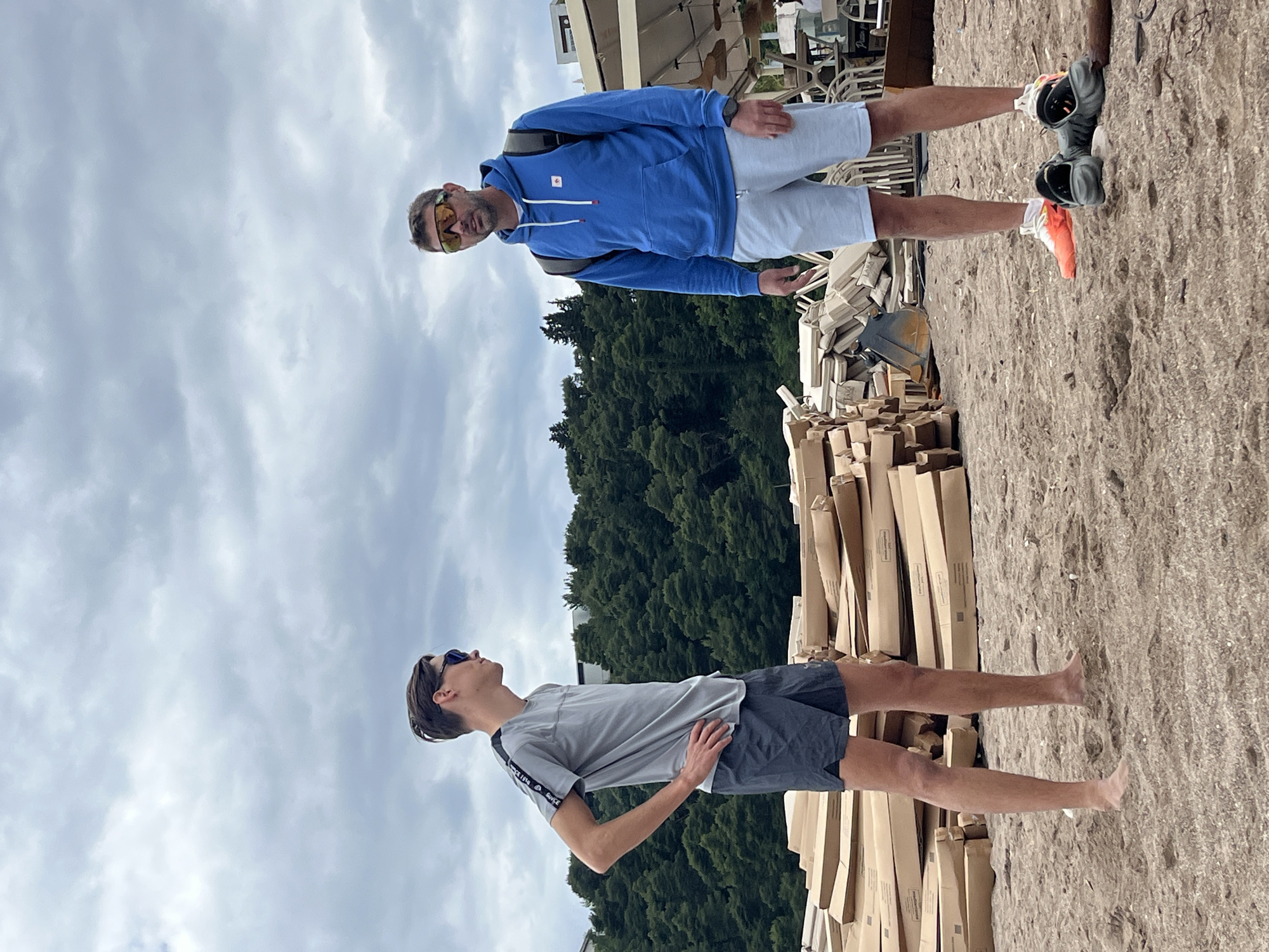 The image depicts two males standing outdoors in Boreti, Montenegro. The foreground shows an area covered with sand, interspersed with a large number of stacked cardboard boxes. The sky overhead is filled with clouds.
The first male (top) is wearing a blue hooded sweatshirt, light gray shorts, and running shoes. He has sunglasses on, is facing the camera, and has a backpack on his shoulders. The second male (bottom) is wearing a gray t-shirt and dark shorts and has sunglasses. He is looking upward and away from the camera. There are green trees visible in the middle ground of the image. A building is seen partially in the background.