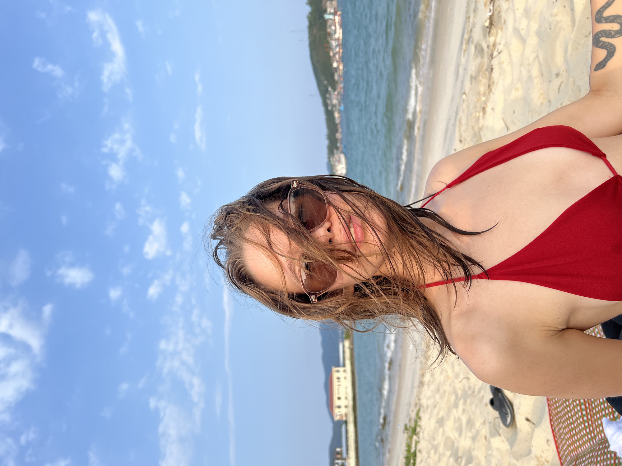 A close-up, slightly off-center portrait captures a young woman on a beach in Nam Nha Trang, Vietnam, on a bright, sunny day. Her wet, medium-brown hair is windblown, partially obscuring her face and the brown-tinted sunglasses she is wearing. A slight smile is visible, conveying a relaxed and happy demeanor. She is wearing a red halter-neck bikini top, and a distinctive tattoo of a winding, snake-like design is visible on her upper right arm.
The background features a clear blue sky with scattered white clouds. Beyond the woman, the calm, blue-green ocean meets a light-colored sandy beach. In the distance, a coastline with lush green hills is visible, dotted with numerous buildings, suggesting an urbanized coastal area. A black sandal and a corner of a colorful patterned mat (red, white, and green) are partially visible on the sand. The image captures a moment of leisure and enjoyment at the beach. There is no visible text in the media.