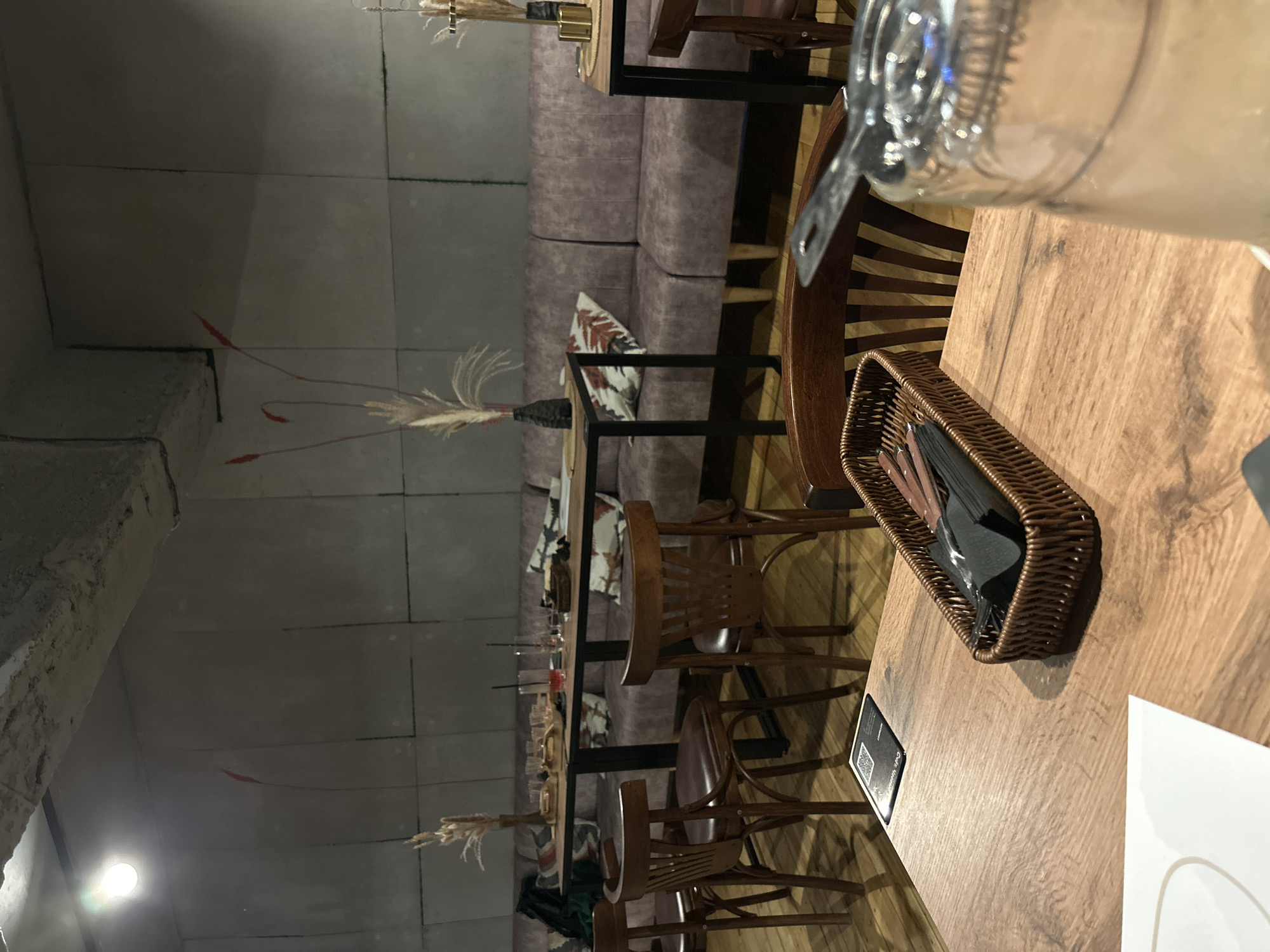 The image shows the interior of a restaurant or cafe with a modern industrial design. The walls are made of concrete blocks, and the ceiling is exposed concrete with visible pipes and lighting fixtures. The furniture includes wooden tables and chairs, and a long plush sofa runs along one wall. Decorative elements include vases with dried grass and minimalist artwork. There are glasses, cutlery in a basket, and a QR code on one of the tables. The lighting is dim, creating a cozy atmosphere. The image was taken in Saint Petersburg, Russia.