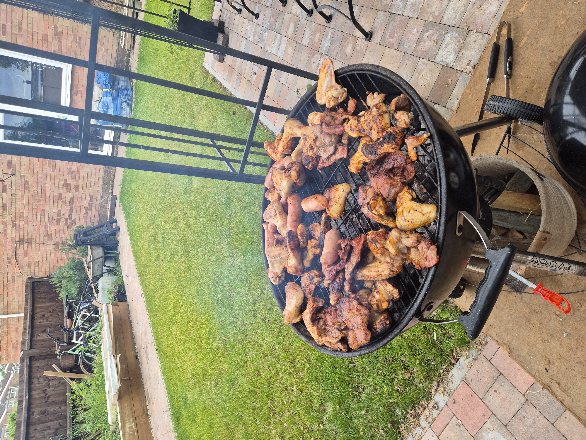 The image shows a barbeque grill filled with cooked meat, including chicken wings, sausages, and other pieces of meat. The grill is located on a patio made of reddish-brown bricks. The lawn is green. There is a trellis, and in the background are a wooden fence, a brick building, and bicycles. The location appears to be a garden in West Suffolk, United Kingdom.