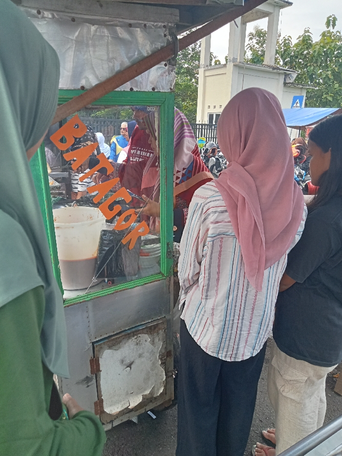 The image depicts an outdoor street food scene in Singaparna, Indonesia, during the daytime, under what appears to be an overcast sky. The central subject is a simple food cart, constructed from metal and glass, around which several people are gathered.
In the immediate foreground, three women are waiting at the cart. On the left, a woman wearing a green hijab is partially visible, facing the vendor. To her right, a woman in a pink hijab and a light-colored striped shirt stands with her back to the viewer, looking towards the cart. Adjacent to her on the far right, another woman with dark hair and a black shirt is also seen from behind.
The glass display section of the food cart prominently features orange letters spelling out "BATAGOR," identifying the type of Indonesian street food sold. Inside the cart, a large plastic pitcher contains a dark liquid, likely a sauce, along with a black plastic bag and other containers. A vendor, partially visible behind the glass and wearing a red garment, appears to be attending to customers.
In the background, a blurred street environment is visible, with other people, some on motorcycles, and a light-colored building with distinct pillars and a tree line under the sky. The scene suggests a casual and busy atmosphere typical of a street food stall catering to passersby.