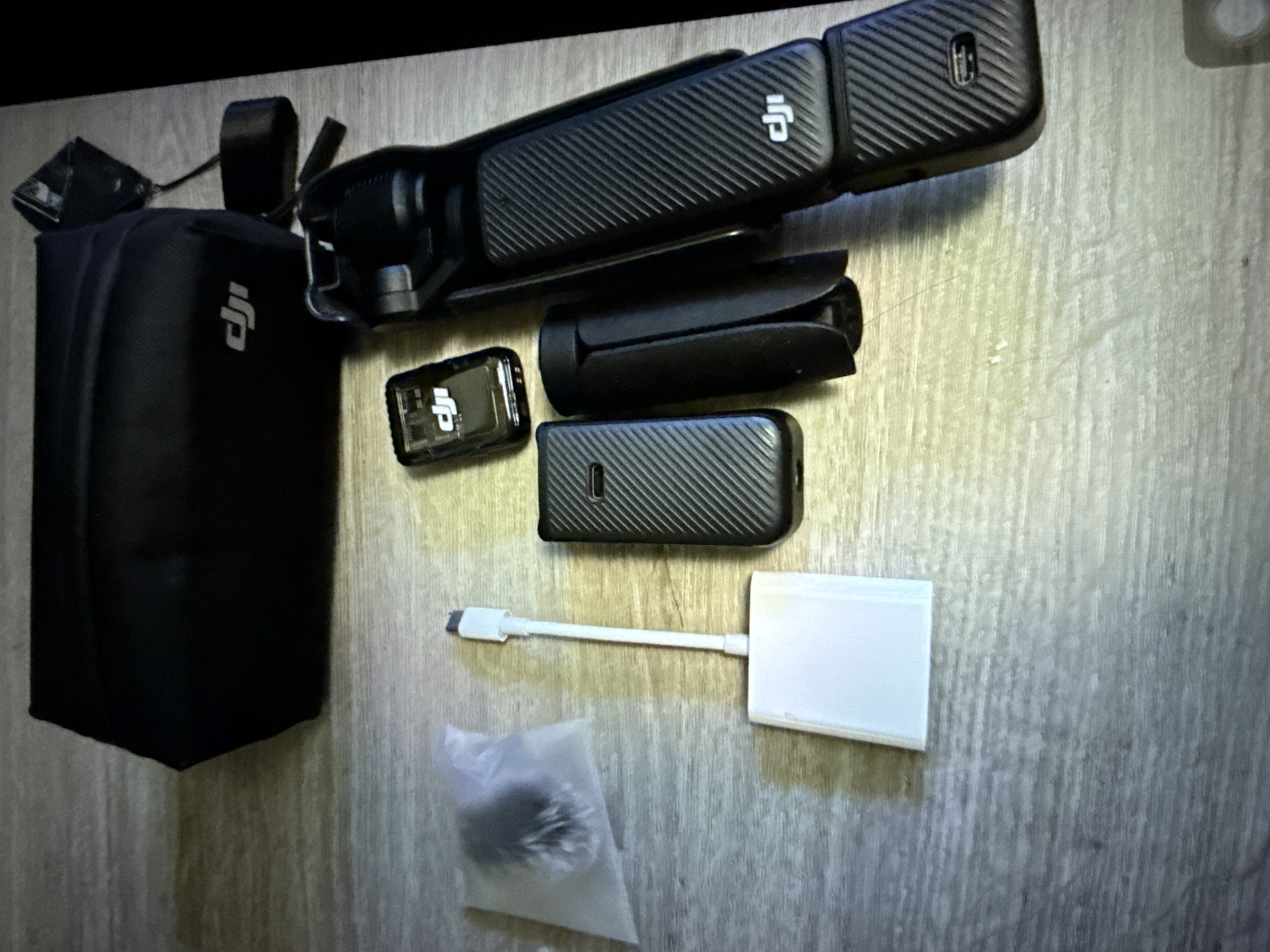 The image shows a collection of black DJI electronic accessories laid out on a light-colored, textured wooden surface. In the top left is a black zippered carrying case with the DJI logo. To its right is a black rectangular accessory attached to a cable. Below the case, to the left, is a small clear plastic bag containing a fuzzy black object. Next to the plastic bag is a white adapter with a white cable. To the right of the adapter is a black rectangular device. Next to that is a black cylindrical object. To the right of these items is a larger black DJI gimbal, which appears to be folded. The gimbal has textured grips and the DJI logo. The image is top-down and illuminated by a warm light source, casting shadows. There are no people present. The scene is not identifiable as being in Taipei, Taiwan or any other specific city.