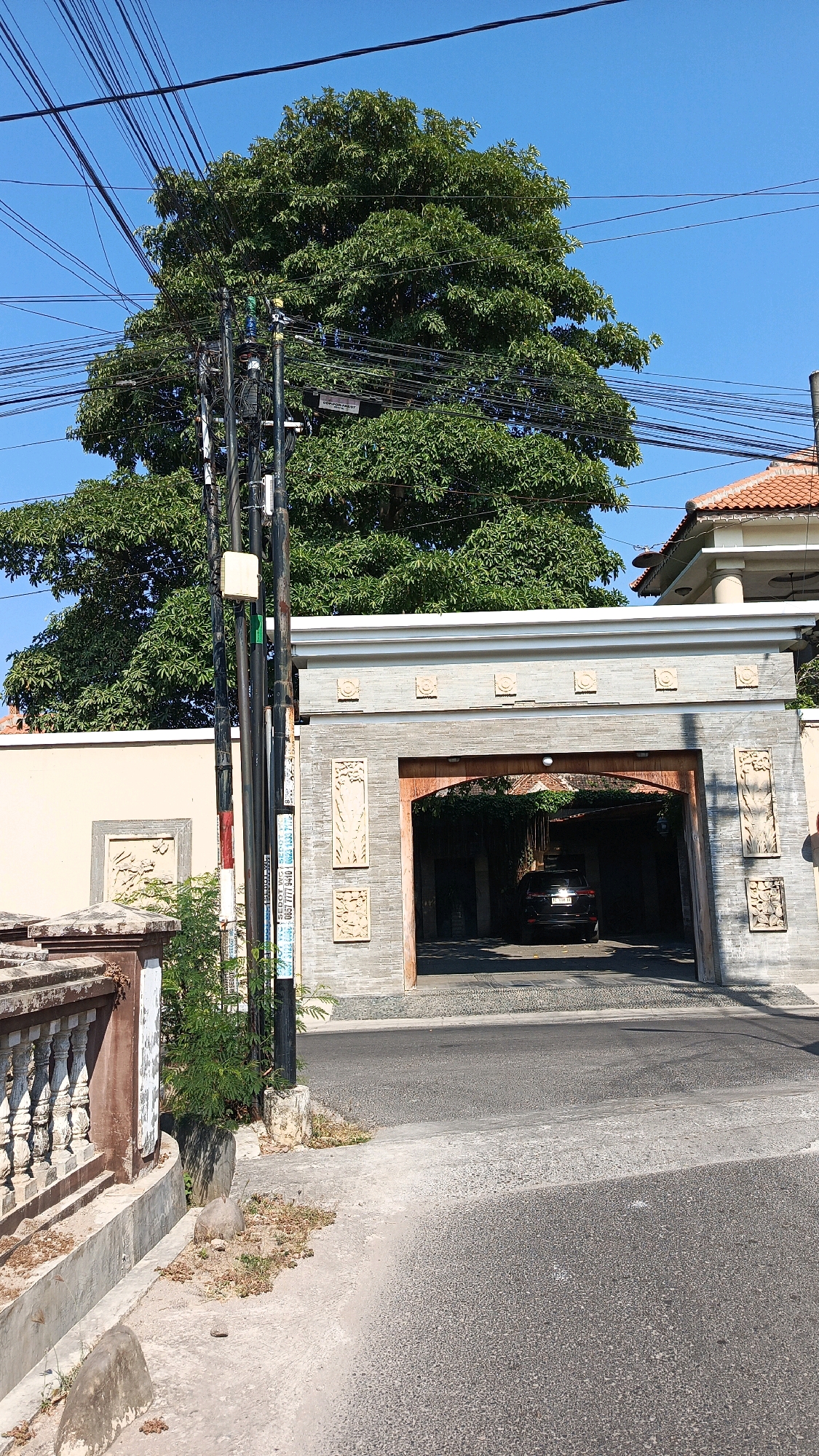 The image depicts an outdoor scene on a clear day, likely in the city of Kertosari, Indonesia, given the presence of a power pole with multiple cables, a common sight in Indonesian cities. In the foreground, a paved road leads towards an arched entryway of a building. The entryway is flanked by decorative stone panels and is constructed with grey stone. A dark-colored SUV is visible inside the entryway. To the left of the road, a concrete barrier with ornate balusters runs along the edge of the property. Behind the entryway and to the left, a tall, mature tree with dense green foliage dominates the upper portion of the frame, its branches intertwined with a complex network of overhead electrical wires. A utility pole with various attachments is situated on the left side of the image, partially obscuring the view of the tree and the building's facade. The overall environment appears to be a residential or commercial area with a mix of natural elements and urban infrastructure.
