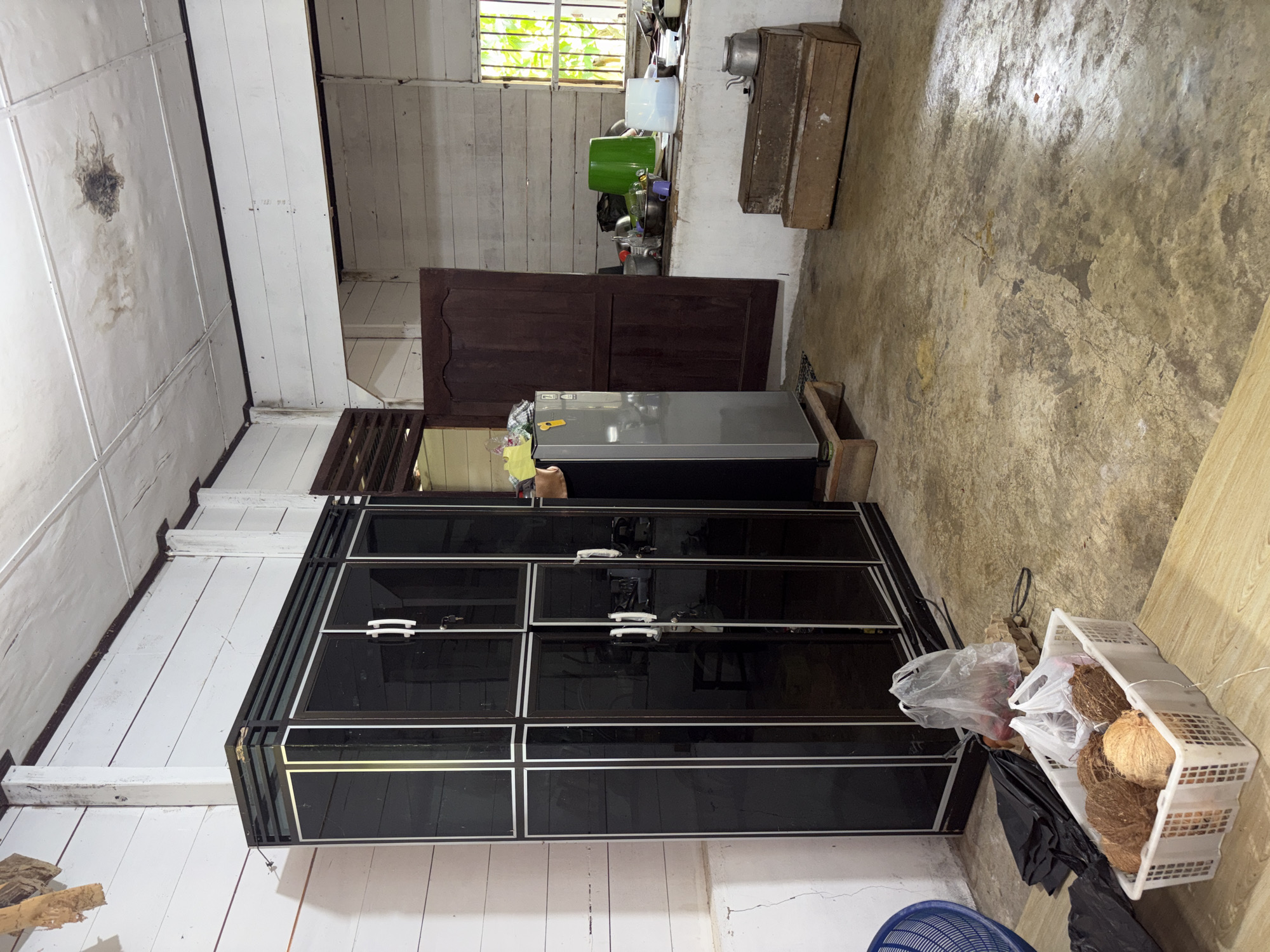The image depicts the interior of a simple dwelling in Beunyot, Indonesia, likely a kitchen or storage area. The room has a worn, concrete floor and walls constructed from horizontal wooden planks, painted white. The ceiling is also made of wooden planks, showing signs of water damage and stains in one area.
Dominating the left side of the frame is a tall, black wardrobe with silver trim and glass doors. Next to it, to the right, stands a grey refrigerator, positioned on wooden blocks. In front of the wardrobe, a white plastic basket contains several coconuts, with plastic bags containing more produce nearby. Black garbage bags are also visible on the floor.
The right side of the room features a wooden door, appearing to lead to another part of the house. Above a low, white counter, a window with metal bars looks out onto green foliage, suggesting an outdoor view. On the counter, various kitchen items are scattered, including bowls, a green plastic bucket, and a white container. Two wooden boxes are stacked on the floor in front of the window, with a metal kettle placed on top of the upper box. The overall impression is one of a functional but sparsely furnished living space.