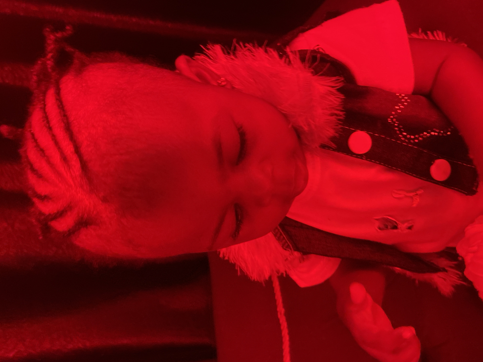 The image shows a close-up of a young child with dark hair styled in braids. The child is wearing a white shirt and a dark vest with fur trim and buttons. The image is saturated with red light, making it difficult to discern fine details and the surrounding environment. The lighting suggests an indoor setting, possibly a home or a studio, but there are no specific landmarks or contextual clues to pinpoint the location within Abakaliki, Nigeria.