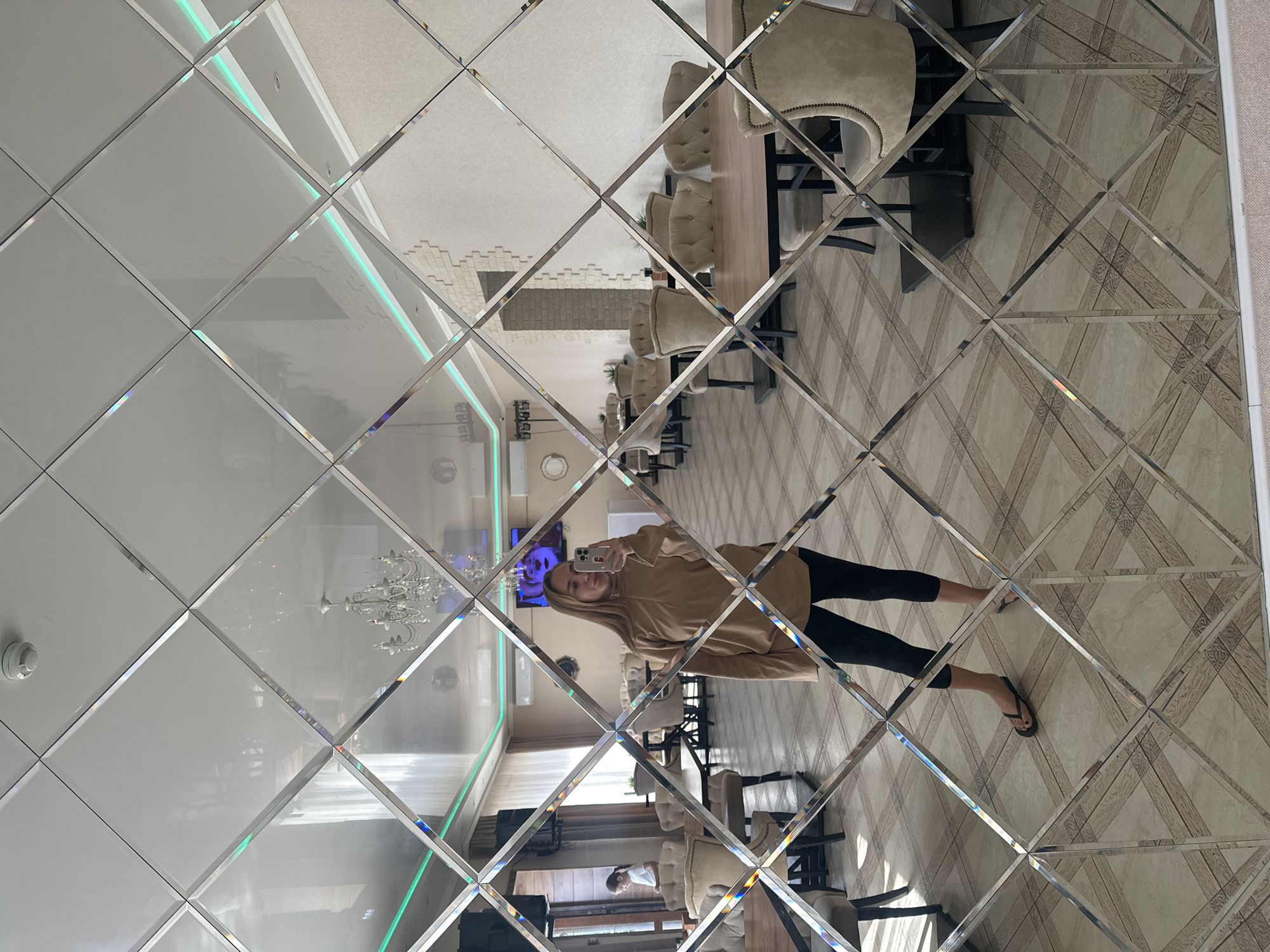 The image shows a reflection in a diamond-patterned mirror wall of a well-lit, modern interior. The reflection reveals a woman taking a selfie with her phone. She is wearing a light brown hooded sweatshirt and black leggings, with black flip-flops. Her hair is long and blonde. The background of the reflection shows a dining area with a long wooden table and several tufted beige upholstered chairs. In the distance, there is a chandelier and some accent lighting on the ceiling. The flooring appears to be a tiled surface with a wood-grain pattern. Another reflection shows a smaller child looking at the camera from further back in the room. The overall impression is one of a contemporary indoor space, possibly a restaurant or a spacious home, during daylight hours. The mirrored surface distorts the reflections, creating a mosaic-like effect.