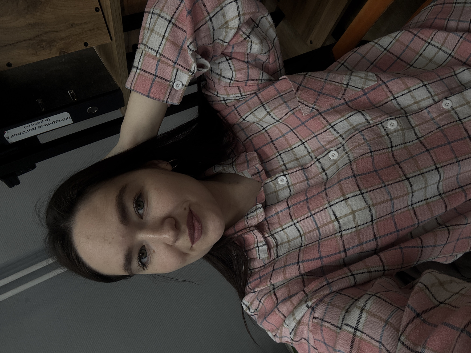 A young woman is depicted indoors, likely in Chaikovsky, Russia, based on the Russian text on a black binder labeled "ПЕРЕДАЧНЫЕ ДОГОВОРА (в работе)". She is wearing a pink and white plaid shirt. The environment includes a wooden surface, possibly a desk, and a grey wall. The woman is looking directly at the camera.