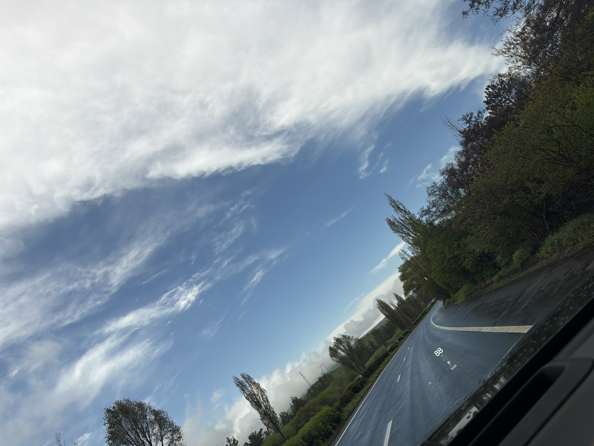 The image is a driver's perspective from inside a vehicle, showing a curved road ahead. The road appears wet, possibly from rain, with a white lane marker visible. Trees and greenery line the side of the road. The sky is partially cloudy with patches of blue visible. The vehicle's heads-up display shows a speed of 88 and the time 12:03. The location is Unknown, Unknown.