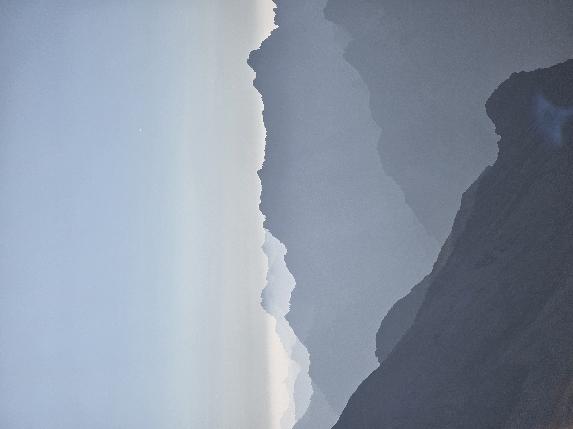 This is a serene, high-angle landscape photograph of layered mountain ranges under a pale blue sky. The foreground features a steep, dark mountain slope with visible texture. As the eye moves towards the background, subsequent mountain ranges appear progressively lighter and more desaturated due to atmospheric perspective, fading into a hazy mist that blankets the mid-ground. The distant peaks are soft, silhouette-like forms against the horizon. The sky is a uniform gradient, starting with a lighter tone near the horizon and becoming a slightly deeper blue towards the top. A faint contrail from an aircraft is visible in the upper right quadrant of the sky. The overall impression is one of vastness and tranquility, with the muted color palette emphasizing the scale of the mountains and the atmospheric conditions. This scene is evocative of the Bavarian Alps near Garmisch-Partenkirchen, Germany, a region known for its dramatic mountain landscapes and frequent atmospheric haze. There are no people, animals, or man-made structures other than the distant aircraft contrail.