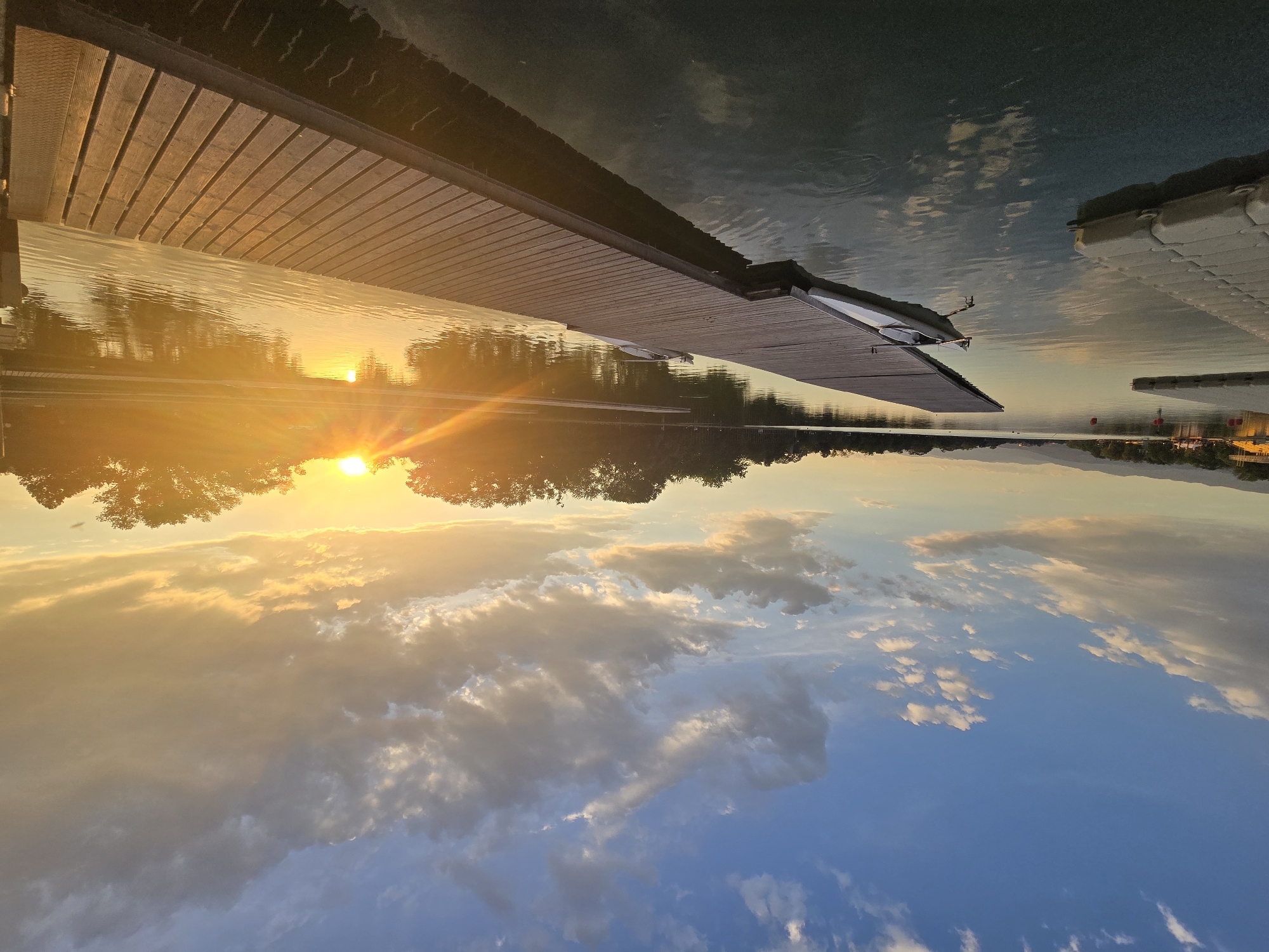 The image depicts a sunset scene on a body of water, likely a lake or large river, at an unknown location. A wooden dock extends into the water from the left side of the frame, with a small boat attached to its end. The sun is setting on the horizon, with its light reflecting in the water and creating a bright glare. Trees line the distant shore, silhouetted against the sky. The sky itself is filled with clouds that are illuminated by the setting sun, creating a gradient of colors from orange and yellow to blue. A secondary, smaller dock is visible in the distance to the right. The photograph appears to have been taken from a low angle, possibly from the water level, and has been rotated.