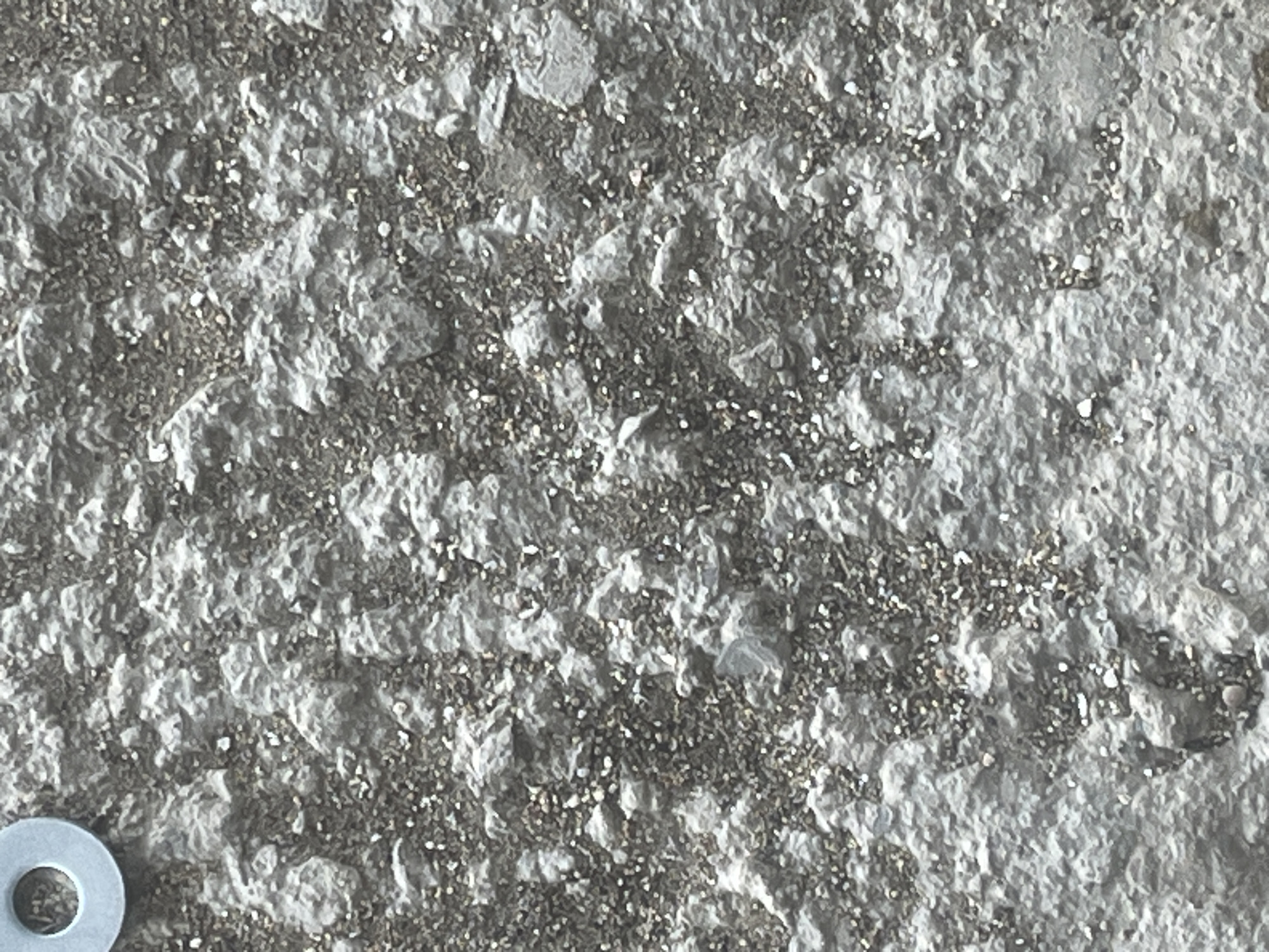The image shows a close-up view of a rough, textured surface, likely concrete or stucco, with small pebbles and debris embedded within it. In the upper left corner, a light blue washer is partially visible. The overall color palette is dominated by grays and whites. The background is out of focus, suggesting the image was taken with a shallow depth of field. No specific location in Kyzylkum, Kazakhstan is identifiable from the image itself.