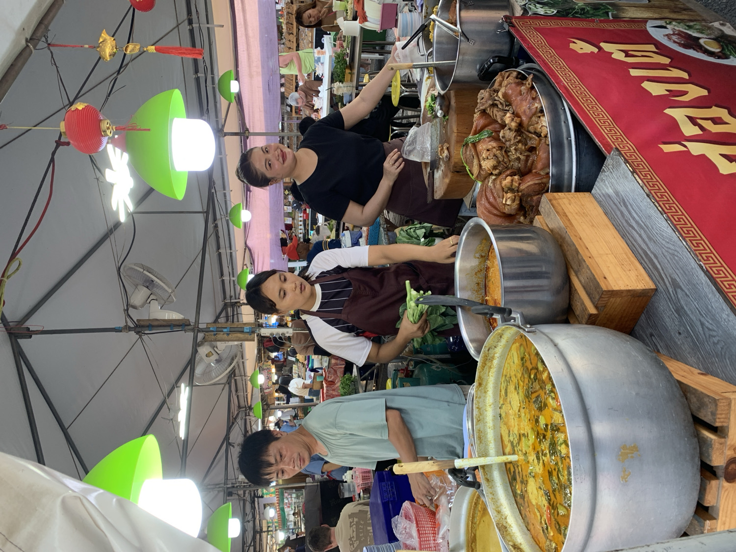 This image depicts a bustling street food stall or market scene in บ้านบางรักษ์, Thailand. Three individuals are actively engaged in preparing and serving food. On the right, a woman in a black shirt and brown apron smiles towards the camera while standing behind a large metal pot filled with stewed pork hocks, characteristic of Thai "Khao Kha Mu." She holds a serving utensil, indicating active service.
Below her, another woman in a striped shirt and brown apron is focused on preparing ingredients, holding green leafy vegetables over a large aluminum pot containing a rich, yellow-orange curry or stew. To the left, a man in a light blue-green shirt is also attending to a similar large pot of curry or soup, with a ladle visible. The pots are large, suggesting a high volume of cooking for many customers.
The stall is set under a white tent or awning structure, from which decorative red lanterns and modern, mushroom-shaped green light fixtures hang, providing bright illumination. Several ceiling fans are also visible overhead, likely for air circulation in the warm climate. In the background, glimpses of other market stalls and people suggest a lively market environment.
A prominent detail is a large red banner with an ornate gold border, displayed vertically on the right side of the stall. Large Thai script is visible on the banner, which appears to read "ขาหมู" (Khao Kha Mu), confirming the stall's specialty as stewed pork hock. The overall scene conveys an authentic and active Thai food market atmosphere during what appears to be daytime, with a friendly vendor at the forefront.