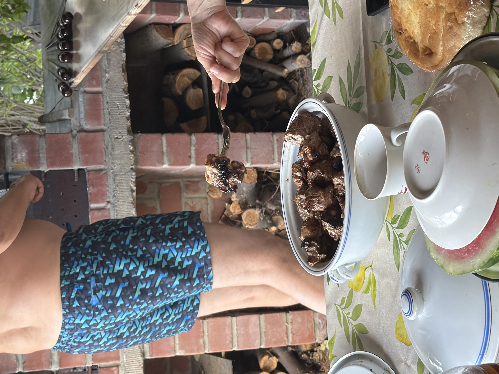 A medium shot captures a person standing next to a brick barbecue pit, likely in an outdoor setting. The person is wearing blue and black patterned swim trunks. In the foreground, a table is set with various dishes, including a large white bowl filled with cooked meat, a covered dish, a slice of watermelon, and a cup. A hand is visible, holding a fork with a piece of meat, suggesting the act of serving or eating. The background shows the barbecue pit filled with logs and a shelf with skewers. The scene appears to be a casual gathering for a meal. The question of the location being Kurgan, Russia, cannot be determined from the visual information present in the image.