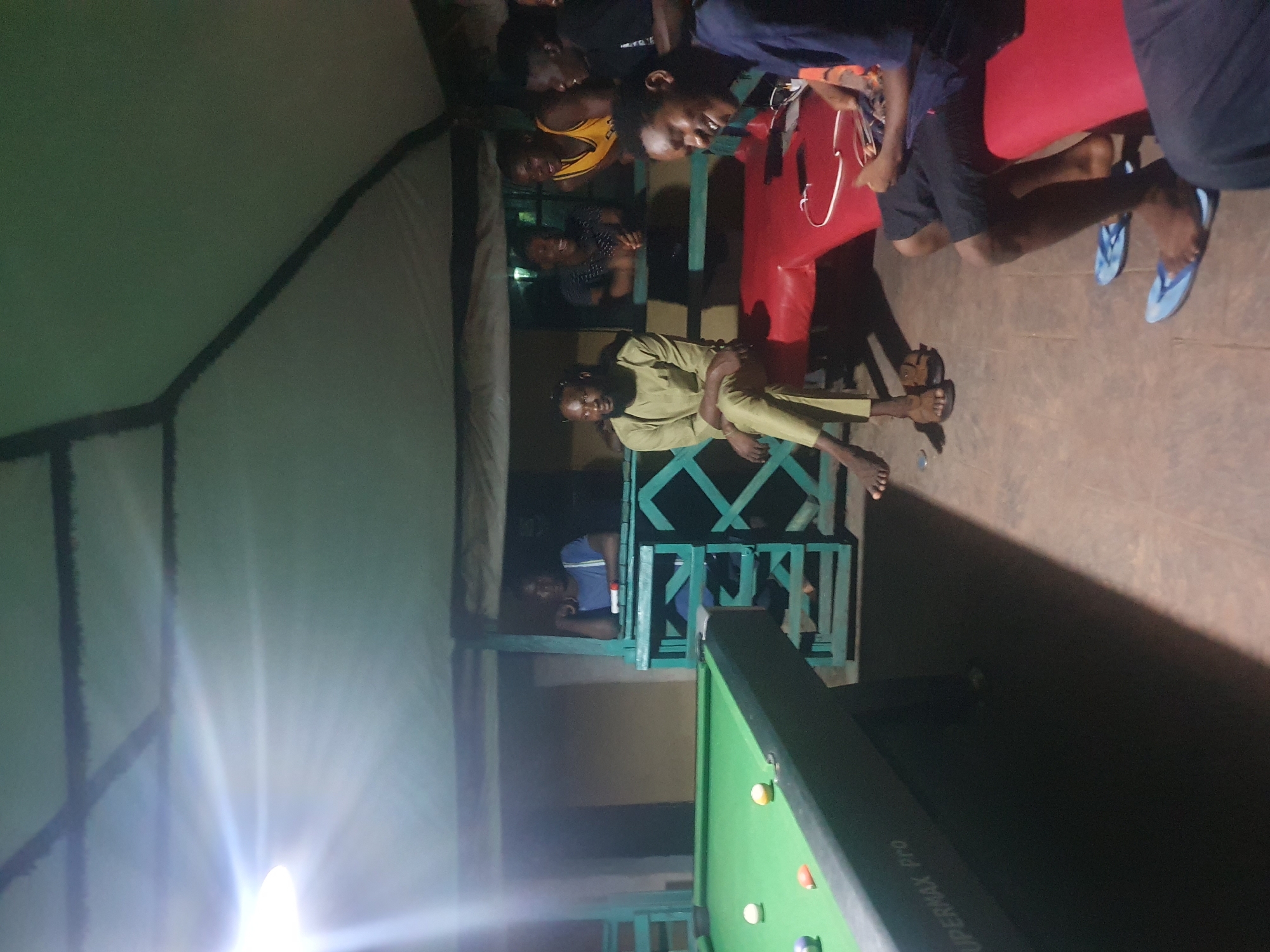 An outdoor pool hall in Lokoja, Nigeria, features a green pool table with several balls, including a white cue ball and colored balls. A group of young men are gathered around, some playing, others observing. The scene is lit by a bright overhead light, creating strong shadows. One man in a light green outfit sits on a red bench, while others stand or lean against a railing. The architecture suggests a simple, open-air structure with a canvas or tarp roof supported by wooden beams. The ground appears to be a dirt or concrete surface.