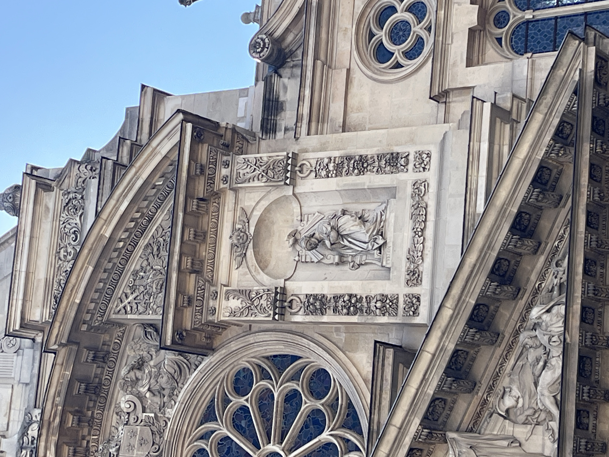 The image shows a close-up, low-angle view of the facade of the Église Saint-Eustache in Paris, France. The facade is made of stone and features intricate architectural details.
Prominently displayed is a large rose window with delicate tracery and blue stained glass. To the right of the rose window, there is a niche containing a sculpture of a robed figure, possibly a religious icon, with a serene expression and hands clasped. Above this niche, there are smaller decorative elements, including winged cherubs.
Further to the right, another smaller rose window with blue stained glass is visible, along with a pointed-arch window below it. The architectural style appears to be Gothic with Renaissance influences, characterized by ornate carvings, moldings, and decorative friezes. The building is bathed in natural sunlight, casting shadows that highlight the sculpted details. The sky above is clear and blue.