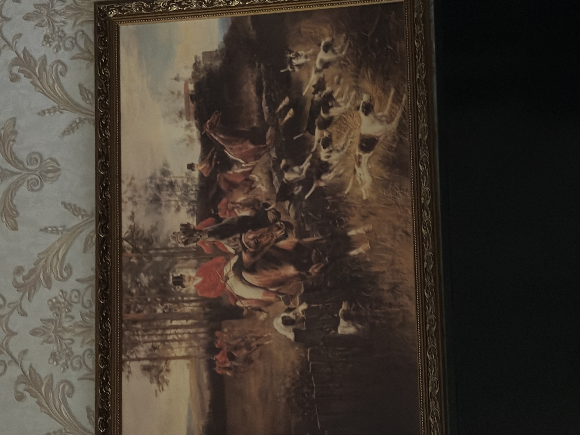 The image is a painting depicting a fox hunt. In the foreground, several dogs are running through tall grass. Behind them, two men on horseback, dressed in red coats and top hats, are also riding. One horse is mid-leap over a fence. In the background, more riders and horses are visible, as well as a wooded area and a building on a hill. The painting is framed in an ornate gold frame. The painting is hanging on a wall with patterned wallpaper. There is no indication of Tambov, Russia in the image.