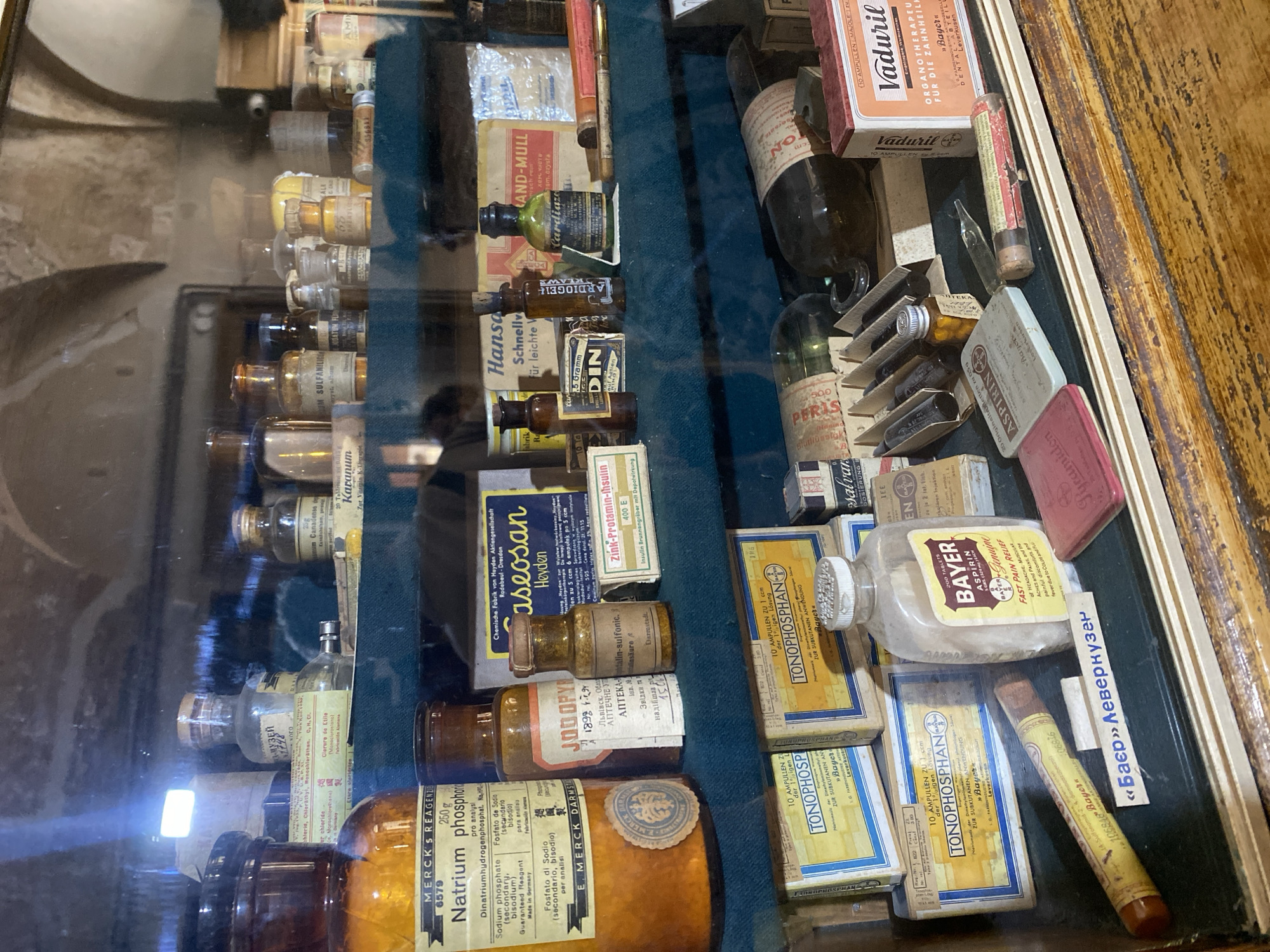 The image shows a display case filled with old pharmaceutical products, likely in a museum or a historical pharmacy setting in Lviv, Ukraine. The items include various glass bottles and vials with paper labels, boxes of medication, and what appear to be ampoules. Some labels are in German, indicating a historical connection to German pharmaceutical companies. Notable items include a large amber glass bottle labeled "MERCK'S REAG... Natrium phosphor", various small bottles with cork stoppers, and boxes of medication labeled "TONOPHOSPHA N", "CASEOSAN", "Zink-Protainin-Insulin", and "VaduriL". There are also small tins, including one labeled "ASPIRIN". The background suggests an interior space with arched architectural features, possibly a historic building. The overall impression is a collection of vintage medical supplies.