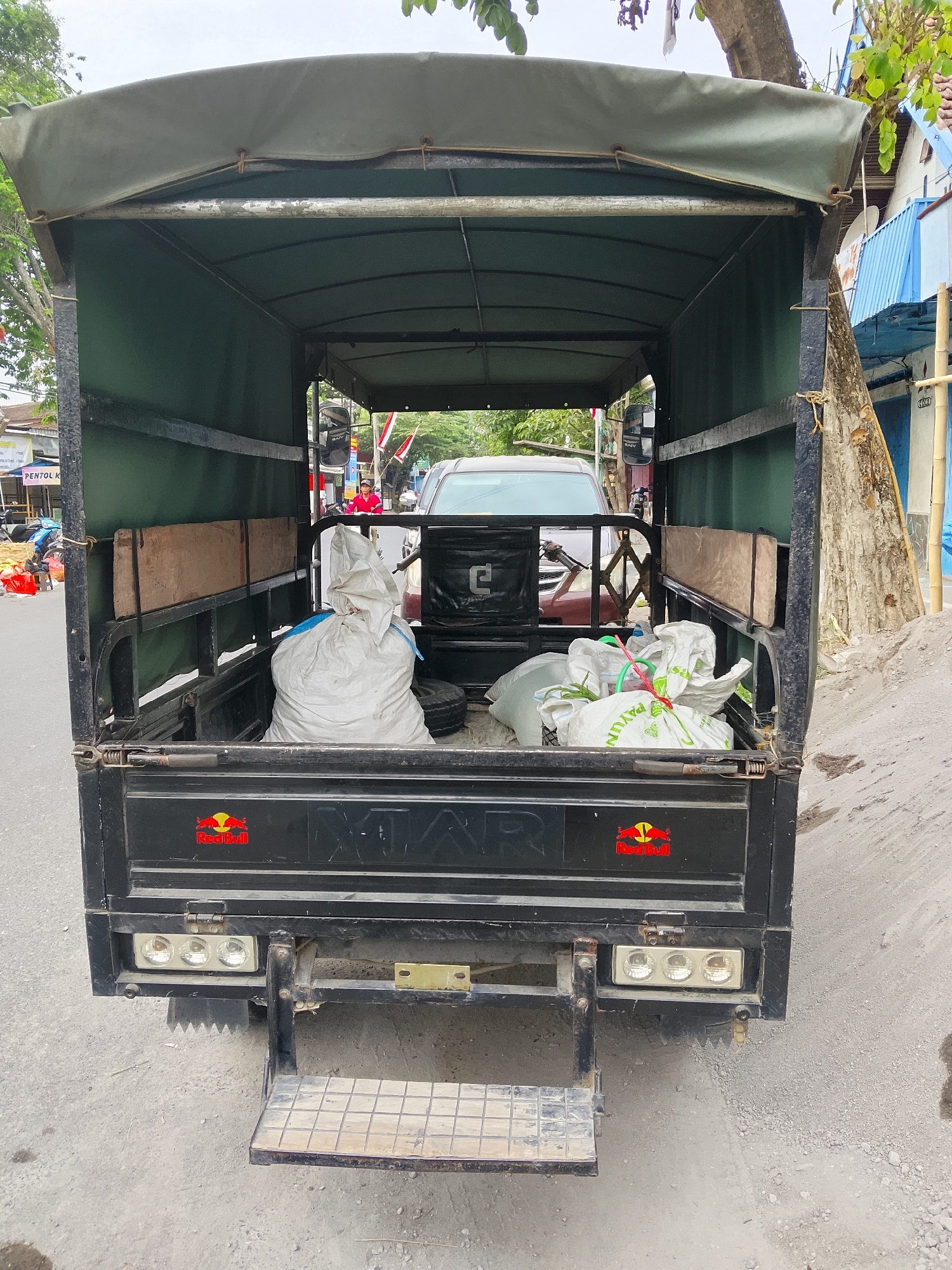 A rear view of a black three-wheeled truck is depicted, parked on a dusty, unpaved road. The truck's bed is covered with a green canvas tarp, supported by a metal frame. Inside the bed, two large white plastic bags are visible, one filled with what appears to be trash, and another containing other items. A spare tire is also present in the truck bed. The rear of the truck features a metal step with a textured surface, and a cluster of three lights mounted horizontally. A red Bull logo sticker is affixed to the back of the truck. The surrounding environment suggests a street scene in Ngelo, Indonesia, with buildings and trees visible in the background, along with other vehicles and people. The overall scene is a snapshot of a functional, utilitarian vehicle in an outdoor urban setting.