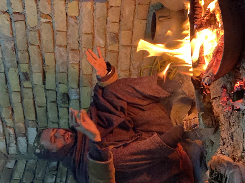 In Kandahar, Afghanistan, a male individual is seated on the ground in a low-light environment, warming his hands over a fire. The man, appearing adult with a beard, wears dark, loose-fitting clothing, a dark shawl, and light-colored athletic shoes. His left hand, with fingers spread, and right hand are extended towards a small fire burning within a shallow metal basin on the ground. Burning wood produces flames and glowing embers. Behind the man and the fire is a textured brick wall made of light-colored bricks. To the right of the fire, a dark metal pot with handles rests on the ground against the wall. Other indistinct objects, including coiled yellow material, are dimly visible in the background.