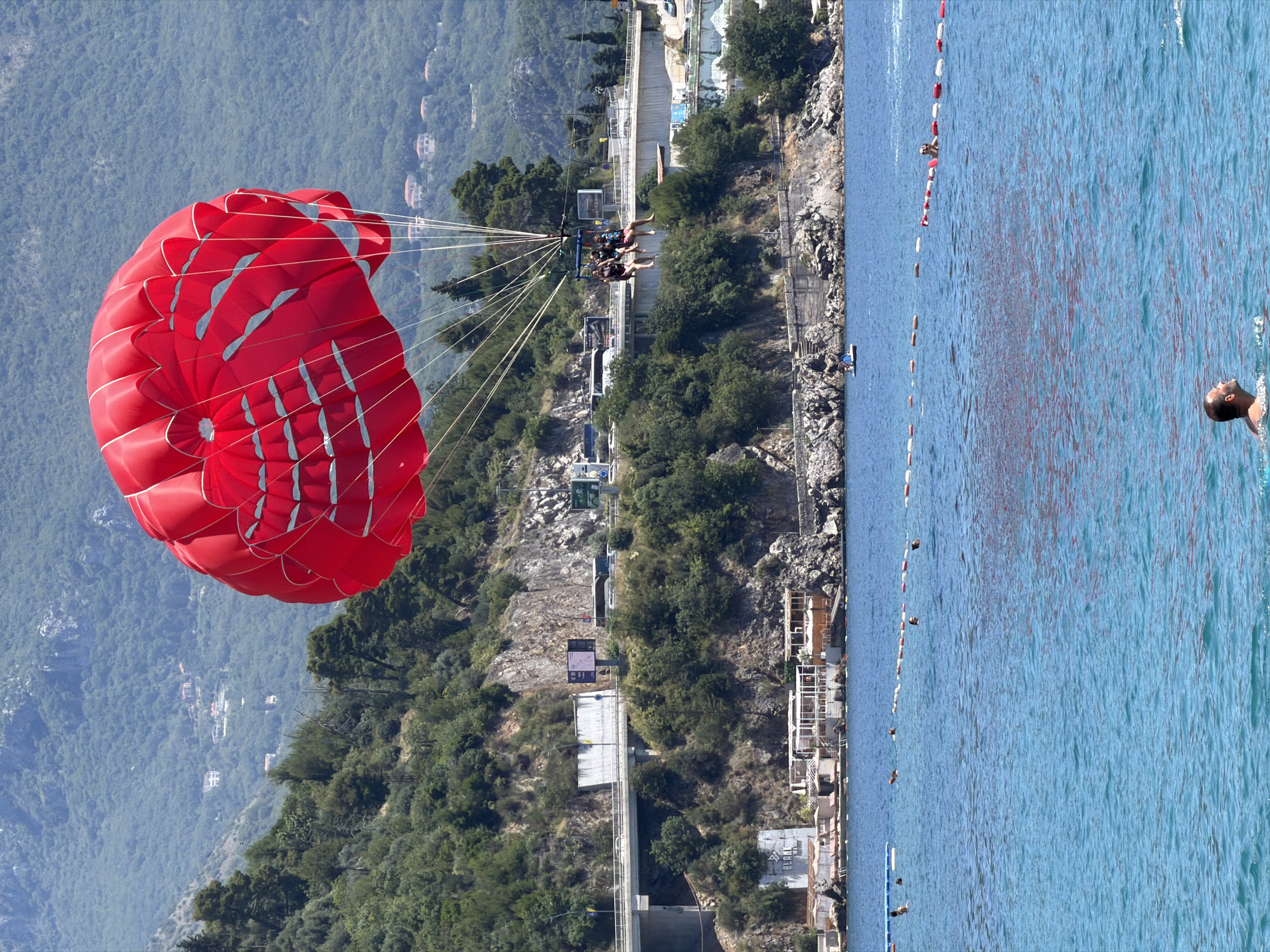 This image captures a vibrant summer scene at a beach in Budva, Montenegro. In the foreground, a man is swimming in the turquoise waters of the Adriatic Sea. In the middle ground, a brightly colored red parachute is carrying two people high above the water, enjoying a parasailing adventure. Behind them, a scenic coastline unfolds with lush green trees clinging to rocky hillsides. A road with vehicles passes along the coast, and a resort or hotel with white structures and umbrellas dots the shoreline. The presence of buoys in the water indicates a designated swimming area. The overall atmosphere is one of recreation and relaxation under a bright, clear sky.