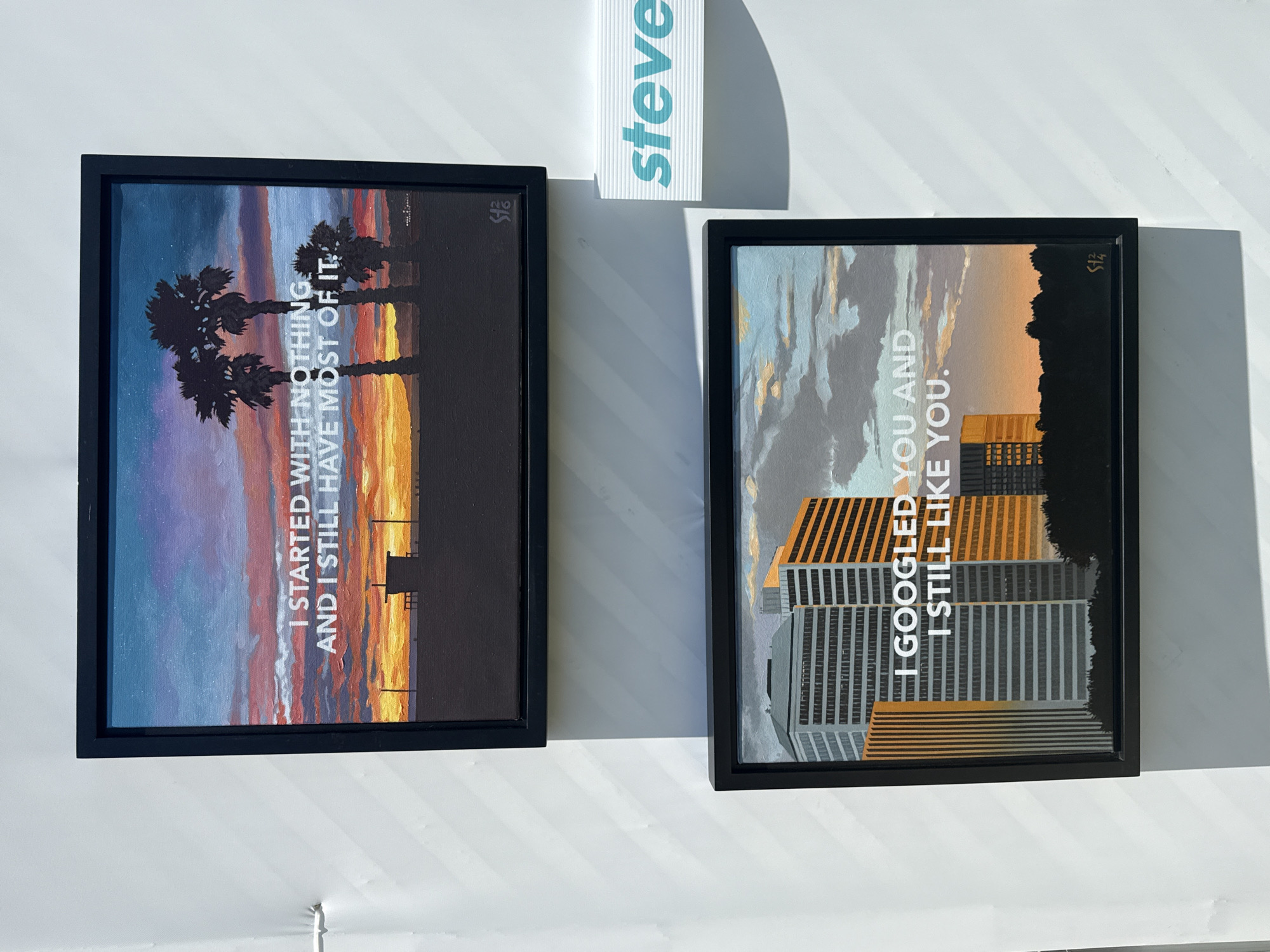 The image shows two framed art pieces hanging on a white wall, possibly in an art gallery or a home. Above the frames, there is a vertical white sign with the name "steve" printed on it in light blue.
The art piece on the left depicts palm trees silhouetted against a sunset sky. White text is superimposed on the image, reading, "I STARTED WITH NOTHING AND I STILL HAVE MOST OF IT." The frame is black.
The art piece on the right shows buildings silhouetted against a sunset sky. White text is superimposed on the image, reading, "I GOOGLED YOU AND I STILL LIKE YOU." This frame is also black.
The lighting in the image indicates a strong light source, causing the surfaces to cast shadows, and the photo appears to be taken indoors. The location is Beverly Hills, United States of America.