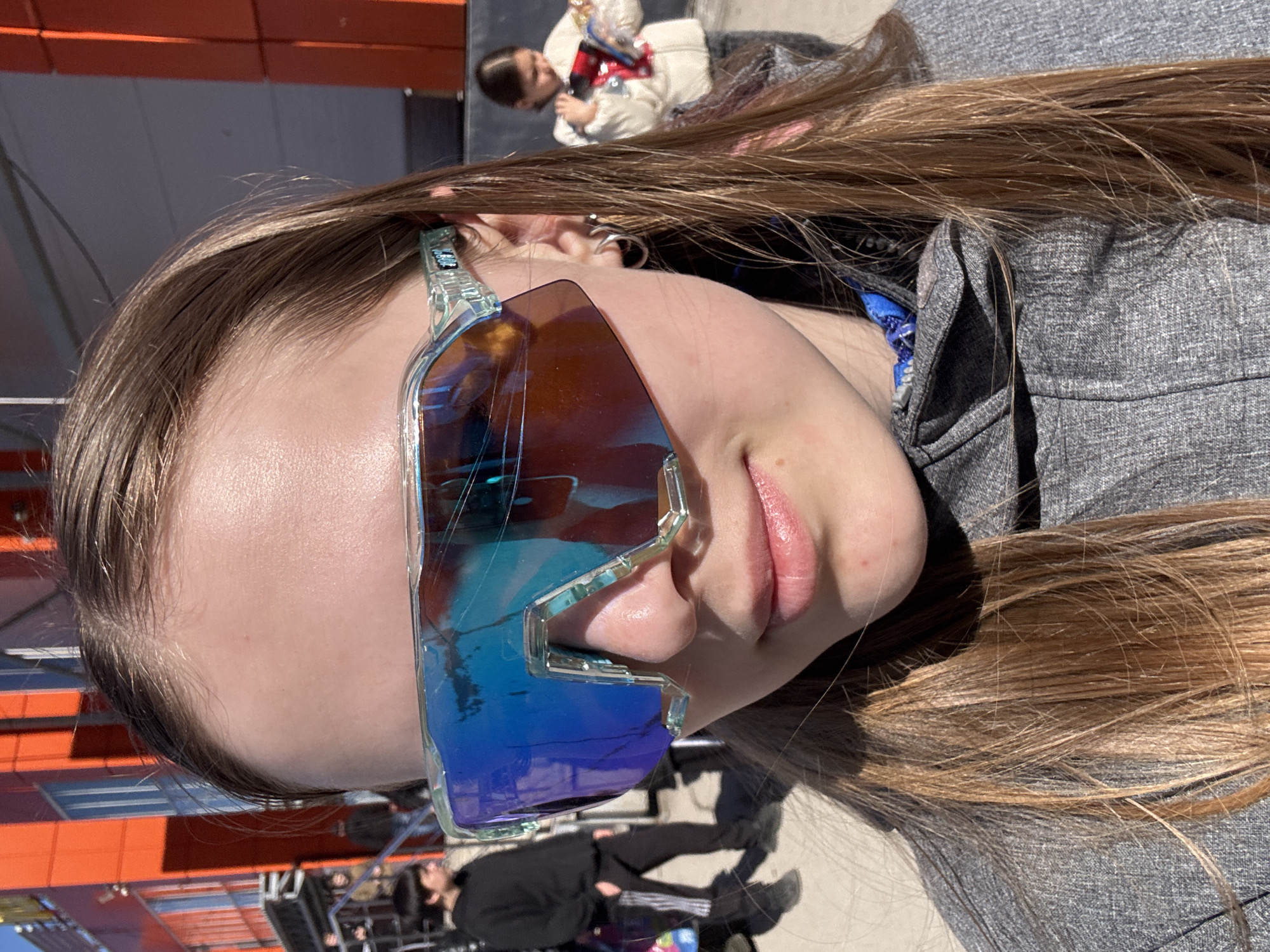 A young woman with long brown hair and light skin is wearing blue-tinted sunglasses with a clear frame. She is wearing a gray zip-up jacket. In the background, a person is seated on a stage, and others are in the foreground. The orange exterior of a building is also visible in the background. The location is likely Usolye, Russia, although specific landmarks are not visible to confirm the exact location.