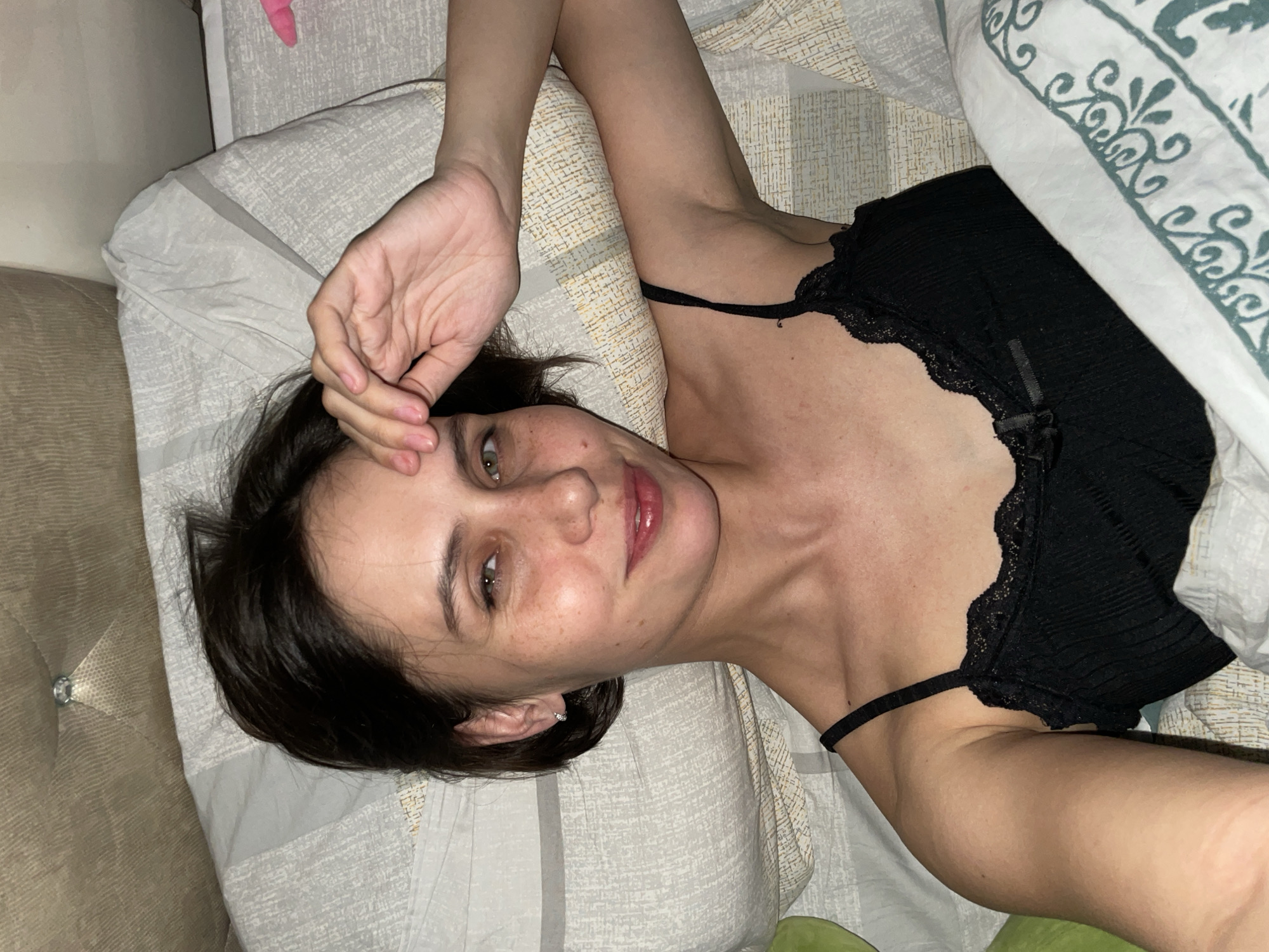 A woman is lying in a bed, wearing a black top with lace trim. Her left hand is raised towards her forehead. She has dark hair and green eyes, and her face has freckles. The bed has patterned sheets and a pillow with a floral design. Part of a tan-colored headboard is visible on the left side. The image does not provide sufficient information to determine the location in Izhevsk, Russia.