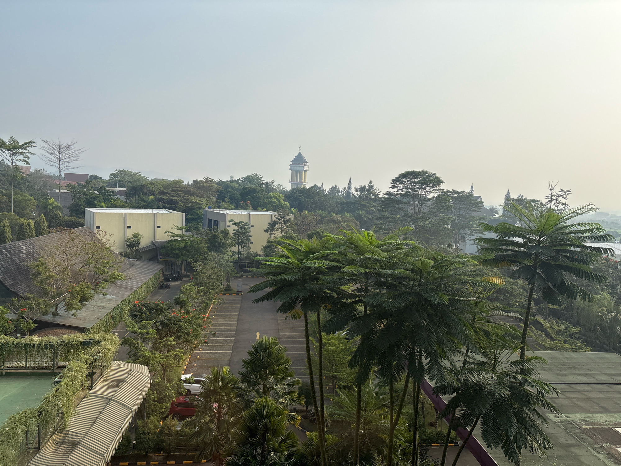 The image depicts a panoramic view of a city area in Pamekaran, Indonesia, likely taken from an elevated vantage point. The scene is dominated by lush green foliage, with numerous trees and palm trees scattered throughout. In the foreground, a parking lot is visible, with several cars parked. Beyond the parking lot, there are buildings with a mix of architectural styles, including a prominent tower with a pointed roof, resembling a minaret. The sky is overcast with a light haze, suggesting either early morning or a slightly polluted atmosphere. The overall impression is one of a tropical urban landscape with a significant presence of natural elements.