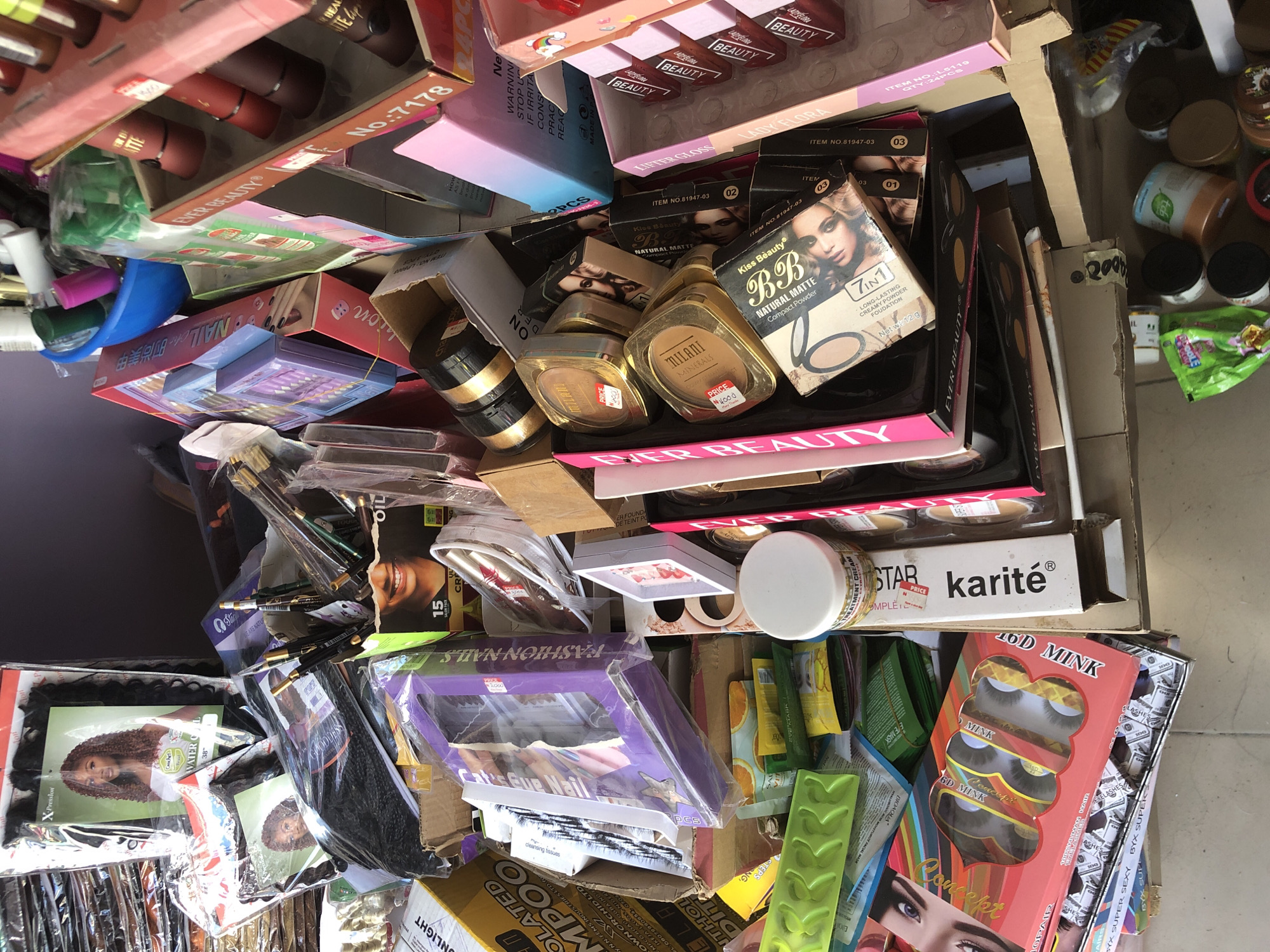 The image depicts a cluttered stall, likely in Ado, Nigeria, filled with an assortment of beauty products. The stall is packed with various makeup items, including lipsticks, powders, eyeshadow palettes, and false eyelashes. Hair extensions are also visible. The products are displayed in boxes and packaging, some of which are stacked precariously. A blue plastic basin containing green and pink items is also present. The overall impression is one of abundance and variety within a small retail space.