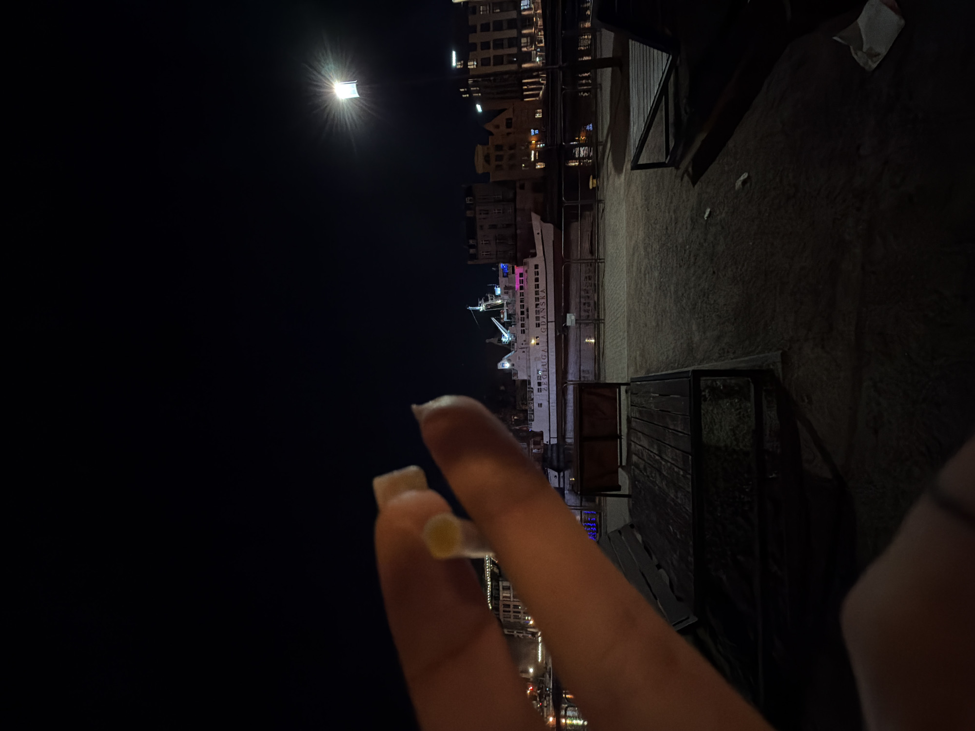 The image is taken at night and shows a hand holding a cigarette. The background includes a waterfront scene in Gdańsk, Poland, featuring buildings and a ship illuminated with lights. The streetlights are visible in the background and there is an overhanging roof in the corner of the image.