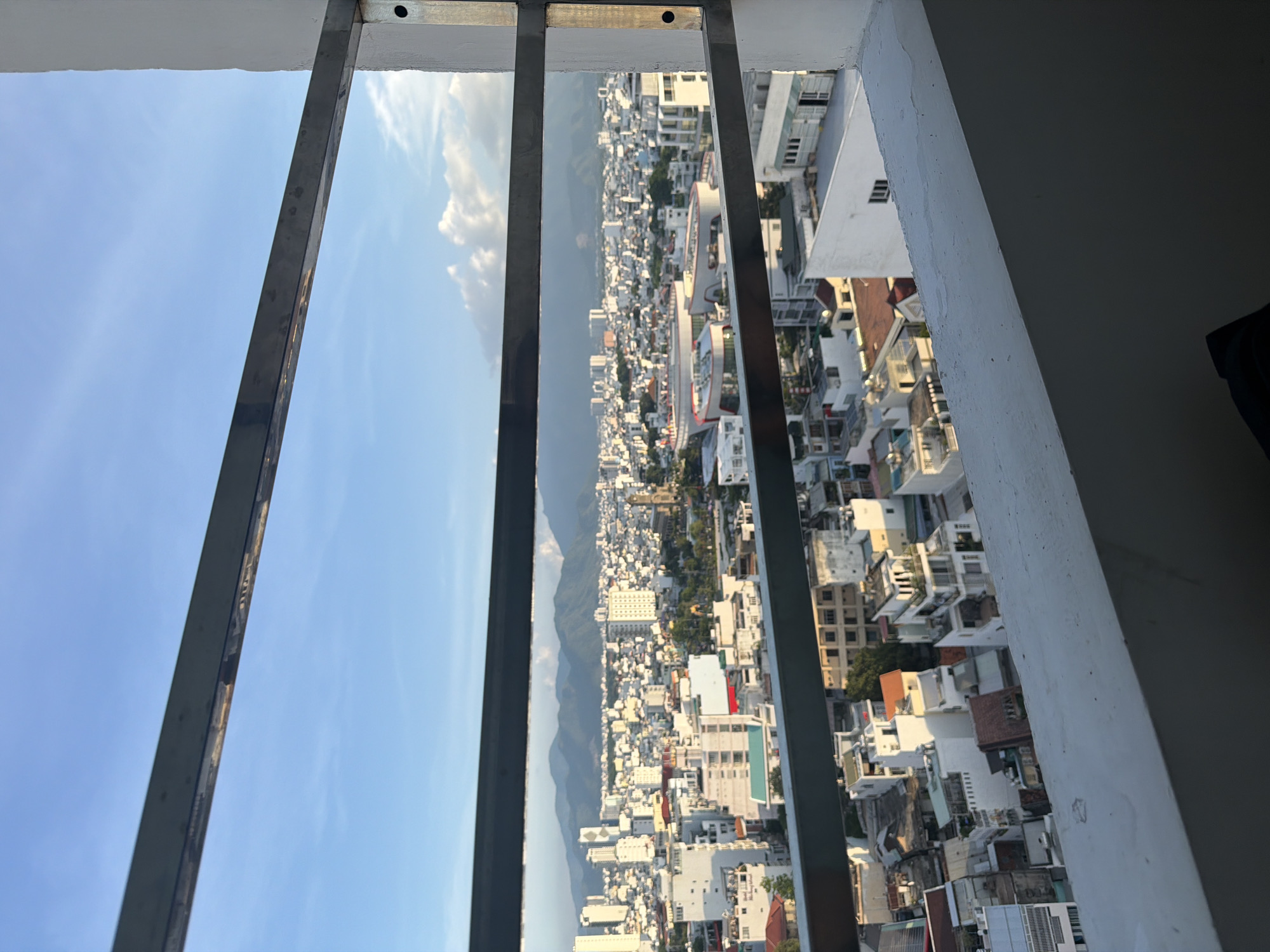 The image depicts a panoramic view of the city of Nha Trang, Vietnam, seen through horizontal metal bars and from a high vantage point, likely a balcony. The city is densely packed with buildings of varying heights and architectural styles, predominantly white and light-colored, suggesting a tropical climate. In the background, rolling green mountains form a natural backdrop under a clear blue sky dotted with scattered white clouds. The perspective is outward, looking towards the horizon, with the foreground dominated by the metallic bars and a concrete railing. The lighting suggests it is either early morning or late afternoon, with sunlight casting shadows and highlighting the building structures. There are no people visible in the frame, only the urban landscape and natural elements.