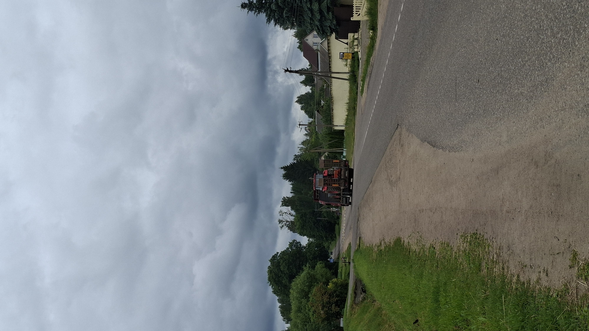 This is a landscape orientation photograph taken outdoors in a rural setting in Belarus. The primary subjects are two red tractors, one in front of the other, on the right side of a paved road. The tractor in the foreground appears to be towing the second tractor. The road runs from the foreground into the middle ground, disappearing behind trees on the left.
The scene is set along a road bordered by a grassy verge and more vegetation. On the left side of the road, there are several houses with light-colored walls and dark roofs, nestled amongst trees. Power lines are visible stretching across the landscape, supported by utility poles. The sky is heavily overcast with dark, stormy-looking clouds.
The time of day is difficult to determine precisely due to the overcast conditions, but it appears to be daytime. The weather is cloudy and potentially rainy or about to rain, contributing to a somber mood.
There is some visible text on a yellow sign attached to the side of one of the houses on the left. The text is small and appears to be in a foreign alphabet, possibly Cyrillic. The overall impression is of a quiet, rural community under a dramatic sky.